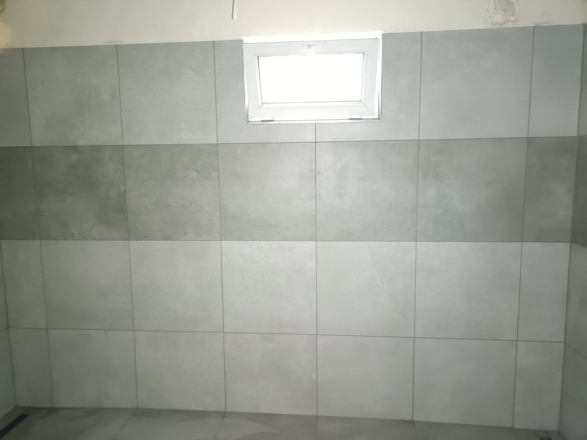 Ściana wyłożona szarymi, matowymi płytkami ceramicznymi, ułożonymi w dwóch rzędach, z oknem w górnej części ściany