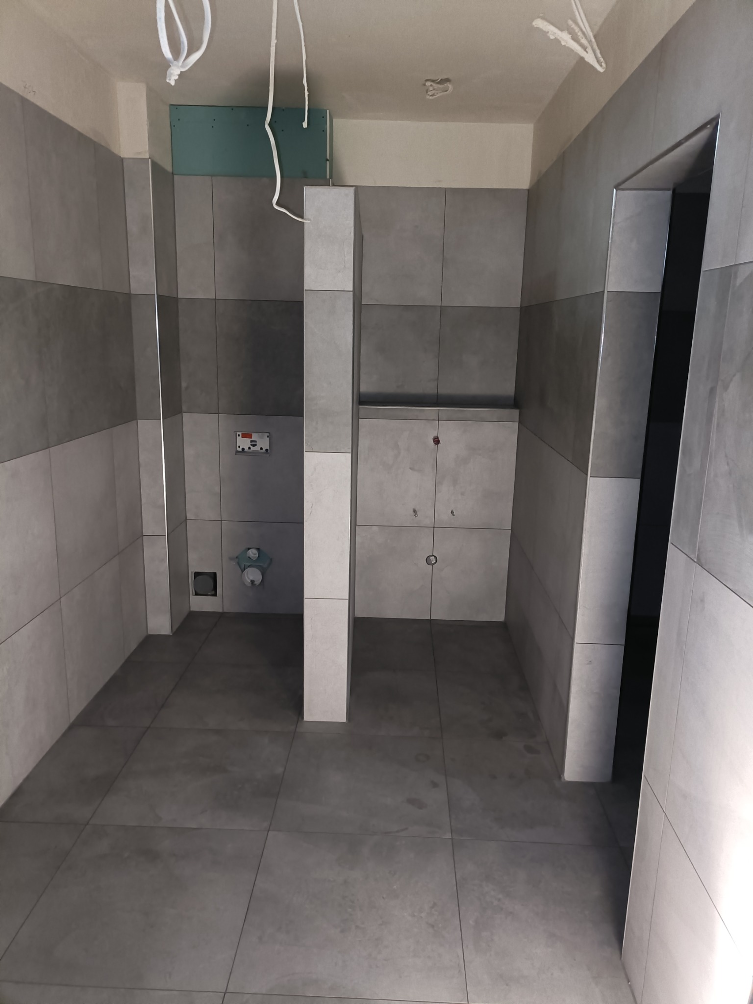 Wnętrze łazienki w trakcie remontu, szare płytki na ścianach i podłodze, widoczne elementy instalacji sanitarnej, sufit z wiszącymi przewodami.