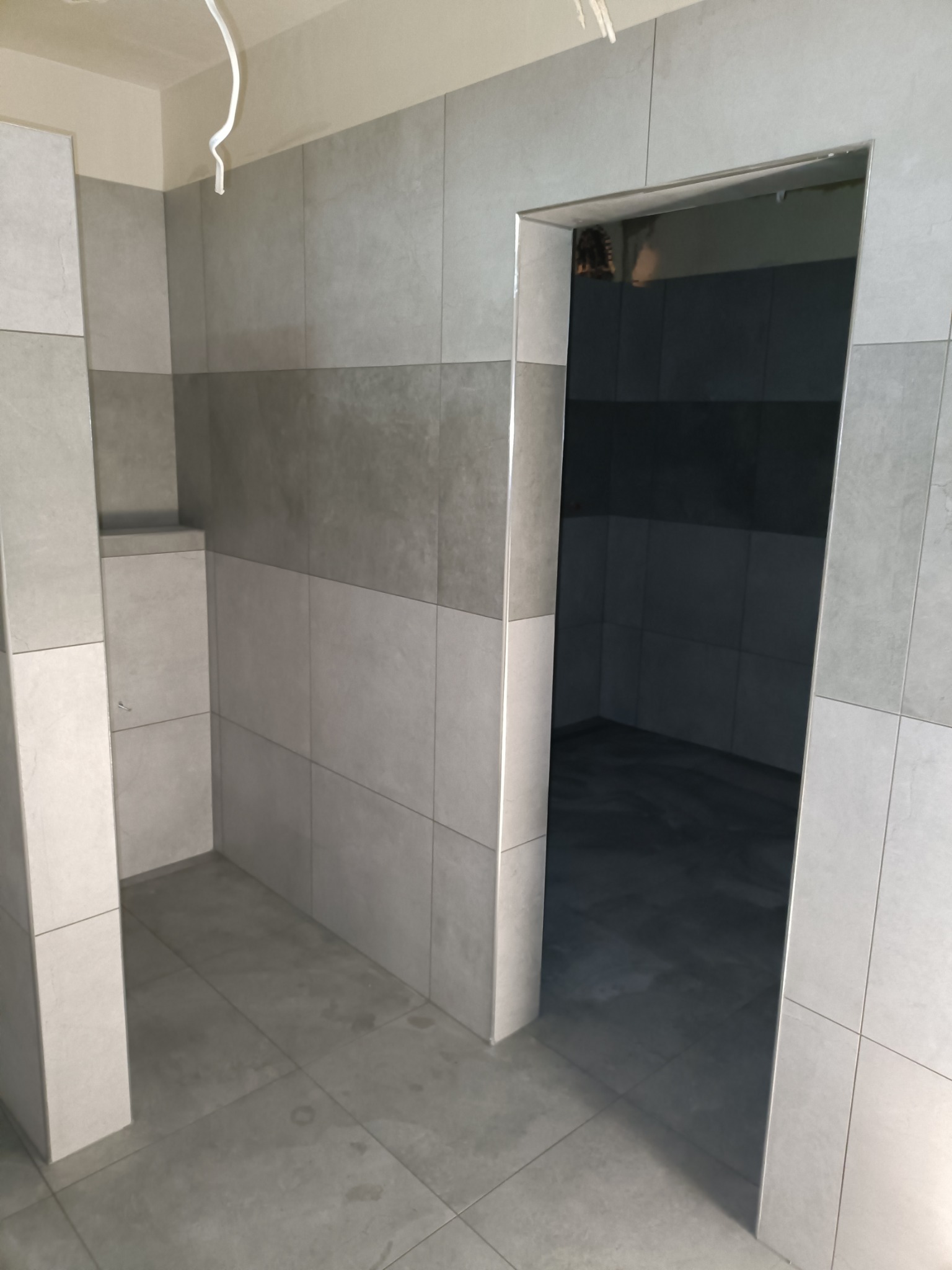 Wykończona łazienka z szarymi, matowymi płytkami ułożonymi w poziomie, widoczne niedoskonałości na podłodze, surowy kabel wystający z sufitu.