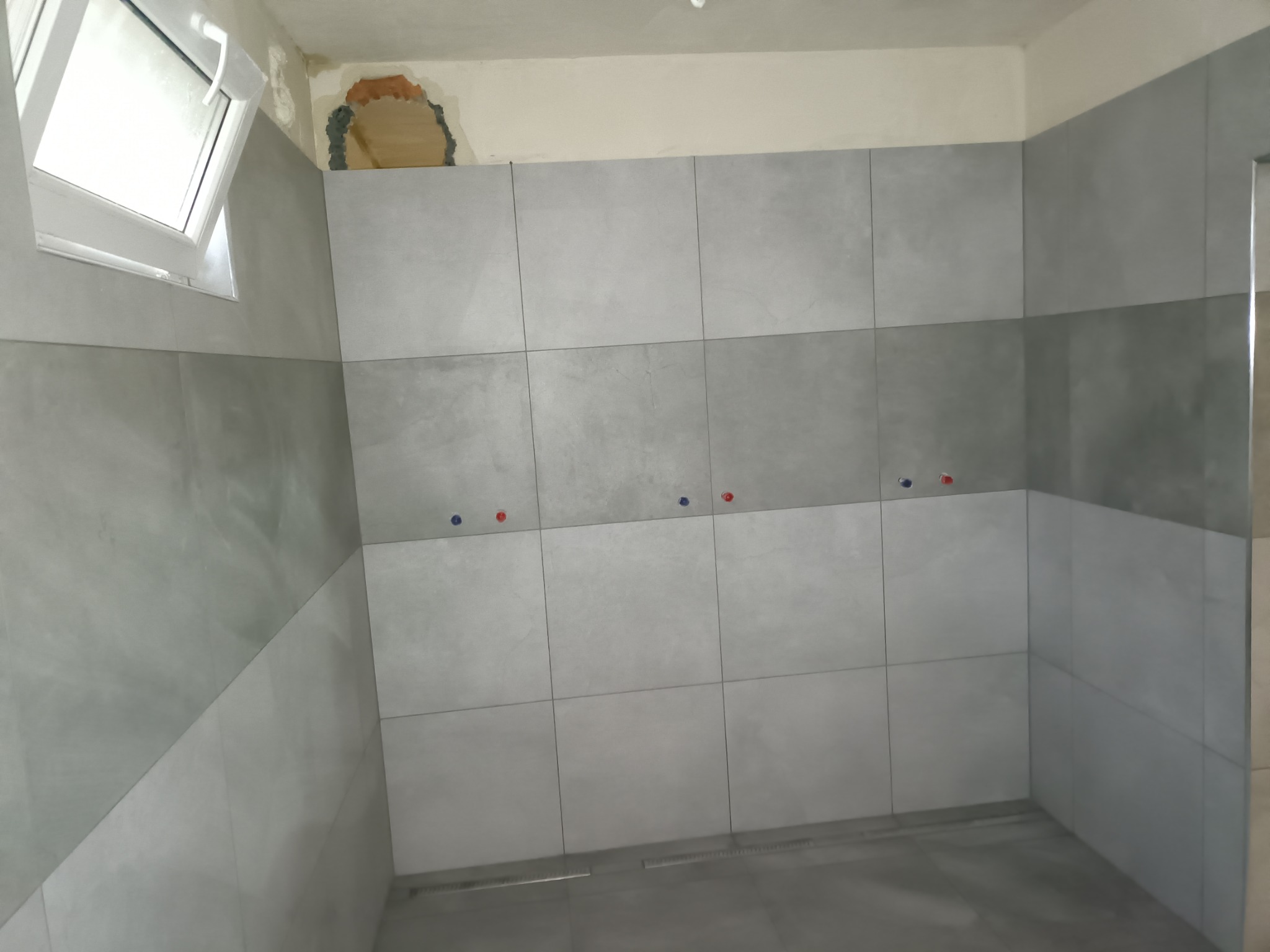 Świeżo wykafelkowana łazienka z szarymi, matowymi płytkami, widoczne otwory na baterie z zaślepkami w kolorach czerwonym i niebieskim, okno dachowe i niedokończony fragment ściany z widocznym tynkiem.