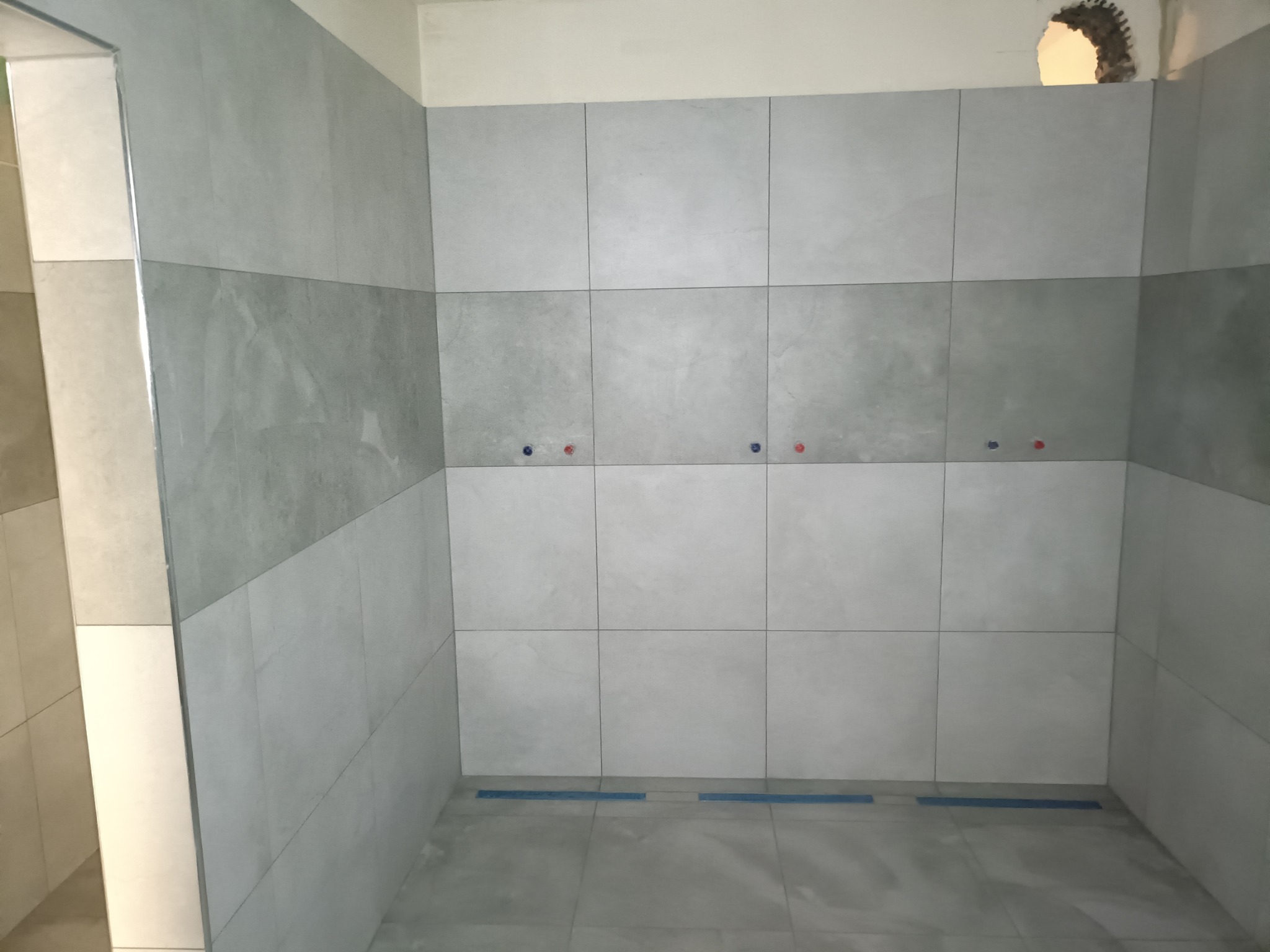 Ściany łazienki wyłożone szarymi płytkami w dwóch odcieniach, z widocznymi przyłączami wodnymi i otworem w górnym rogu ściany.