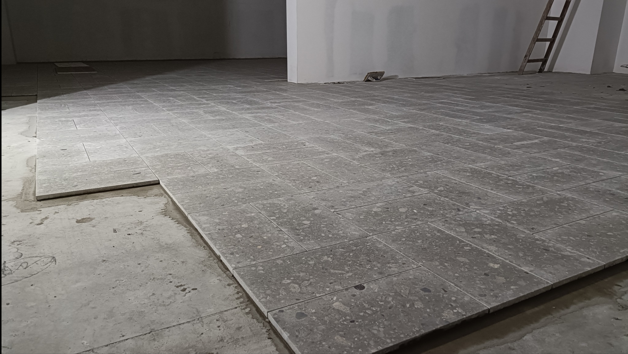 Układanie szarych, prostokątnych płytek podłogowych w jodełkę na surowym, betonowym podłożu, widoczne niedokończone fragmenty i drabina oparta o ścianę.