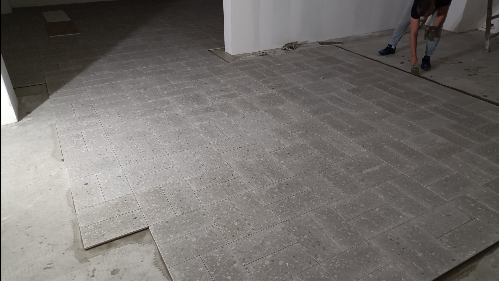 Układanie szarej, drobnoziarnistej terakoty w jodełkę na podłodze w trakcie prac wykończeniowych; widoczny fragment ściany i osoba dociskająca płytki.