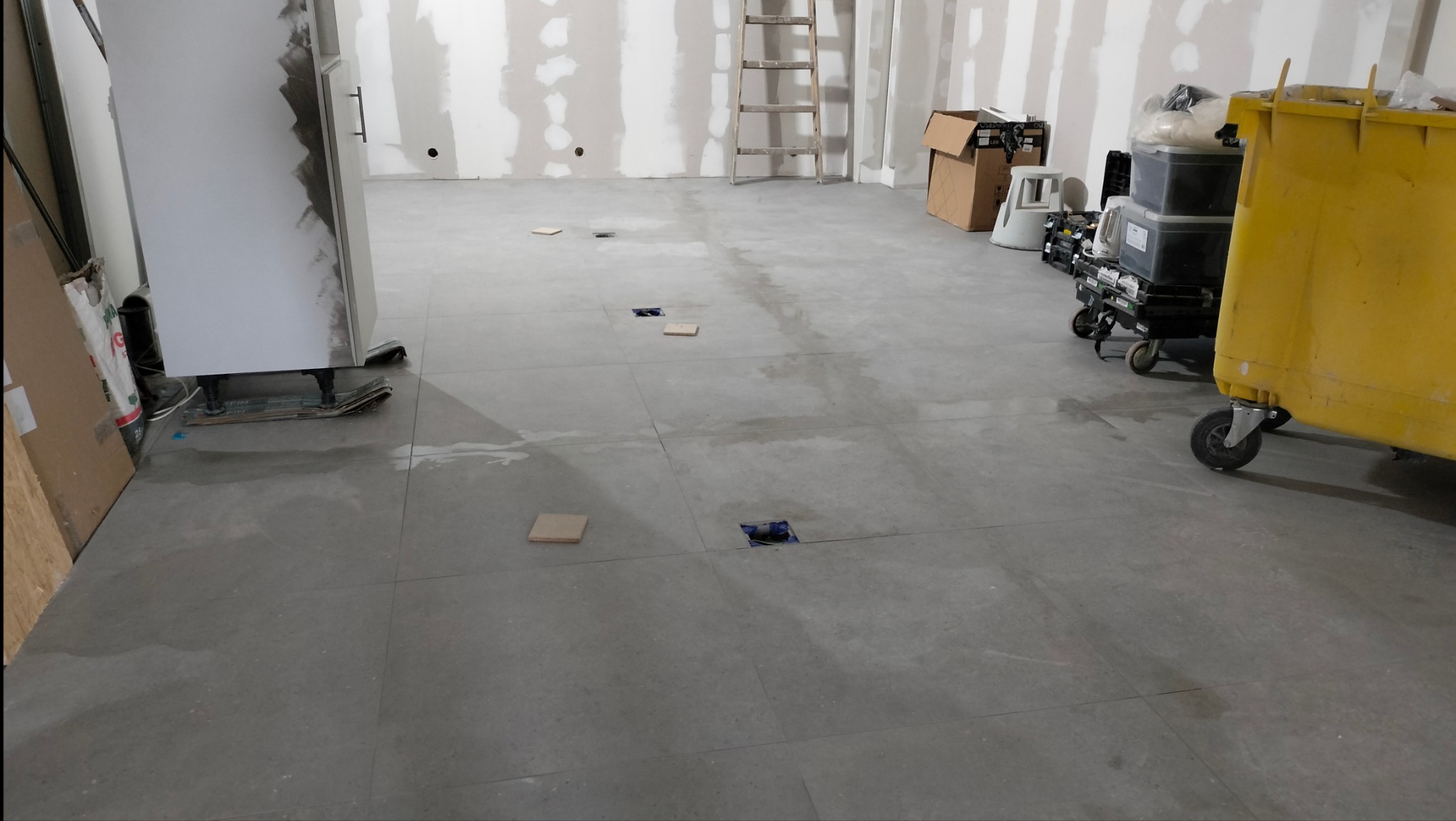 Szare płytki podłogowe układane w dużym pomieszczeniu, widoczne otwory na instalacje z tymczasowymi przykryciami, ślady po zaprawie, drabina oparta o ścianę, pojemnik na odpady budowlane.