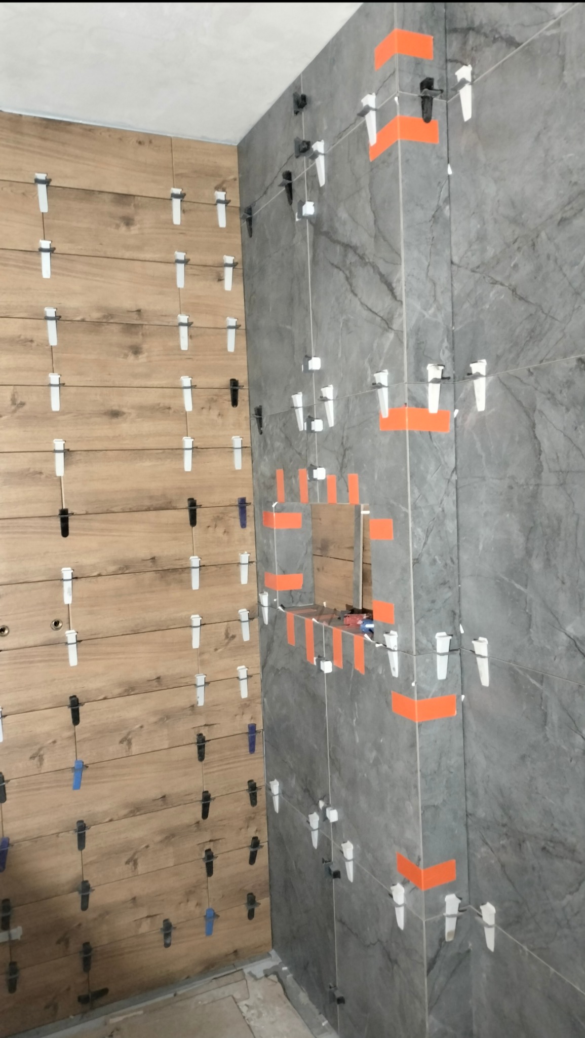 Ściany wyłożone płytkami imitującymi drewno i marmur, z użyciem systemu poziomowania płytek, widoczne pomarańczowe taśmy maskujące.