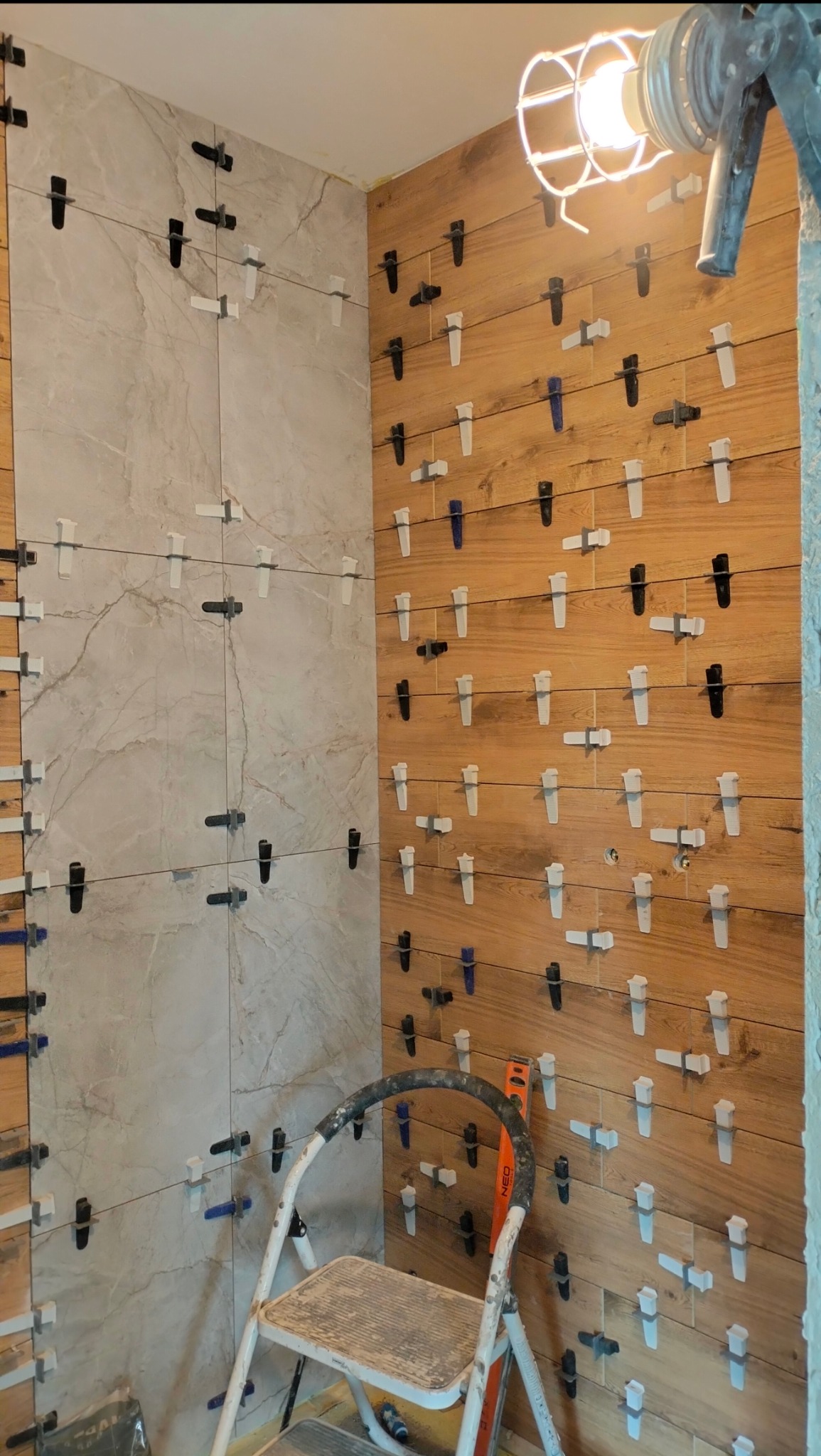 Układanie płytek imitujących drewno i marmur na ścianie z użyciem systemu poziomowania, widoczna drabina i lampa budowlana.
