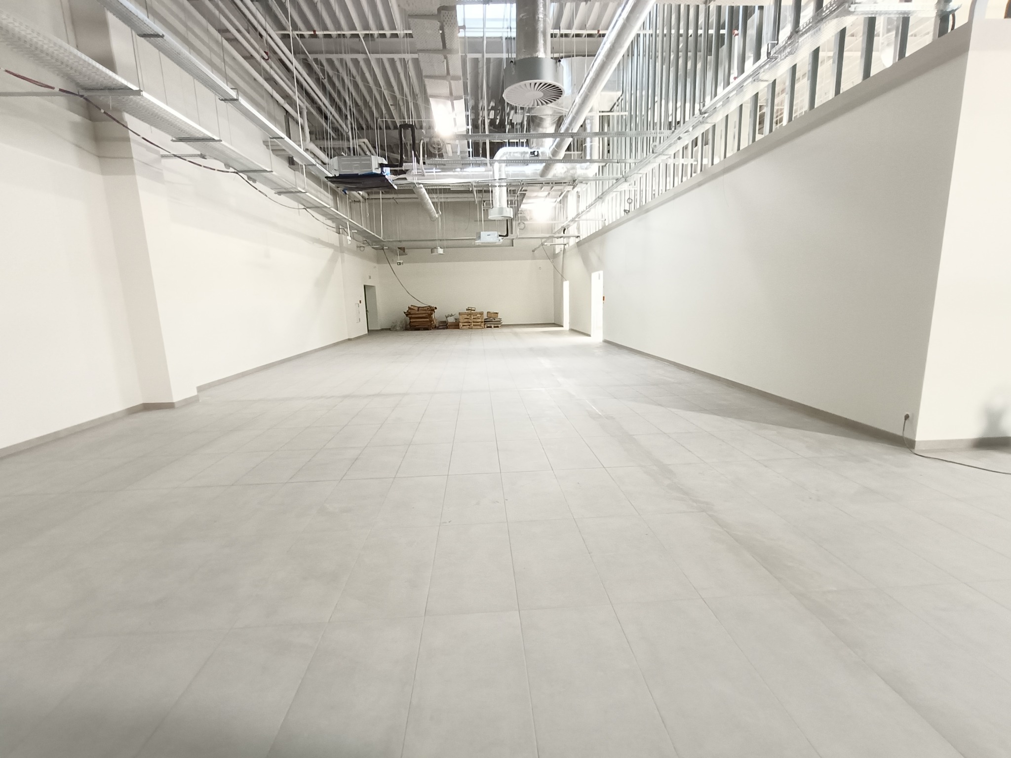 Wykończone wnętrze komercyjne z jasnoszarą podłogą z płytek, widoczne instalacje pod sufitem i palety z materiałami w oddali. Minimalistyczny, nowoczesny design.