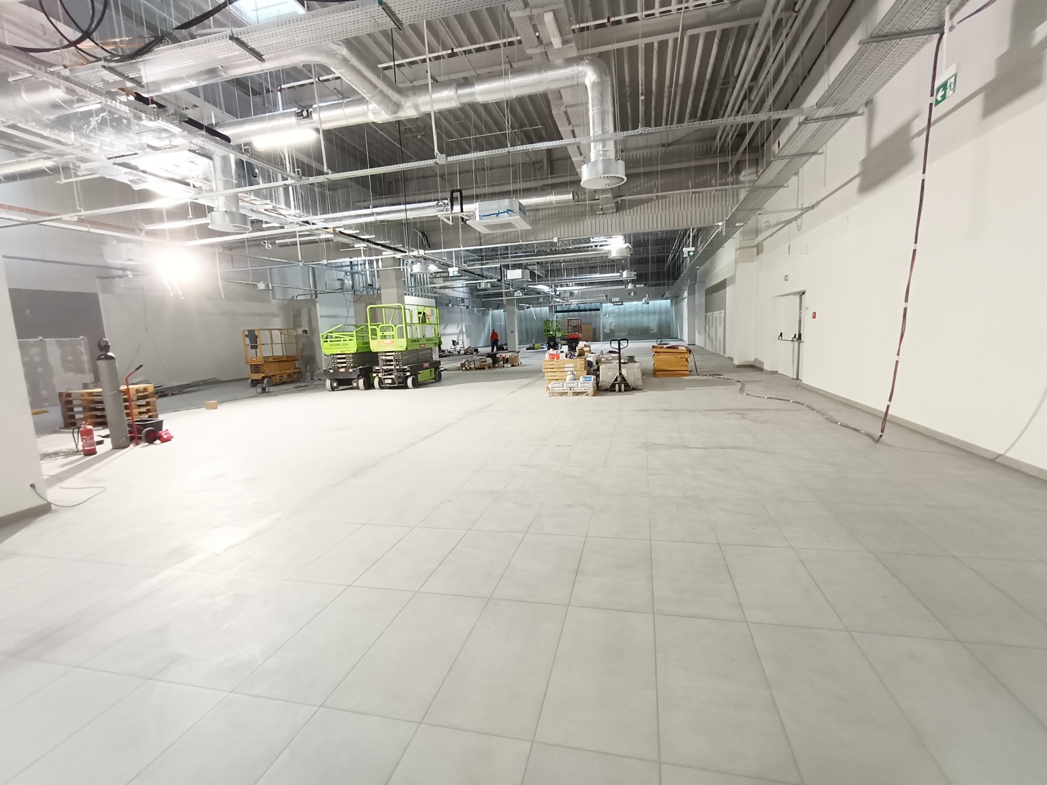 Jasne, przestronne wnętrze hali z ułożoną szarą podłogą z płytek. Widoczne maszyny budowlane, palety z materiałami i instalacje pod sufitem.