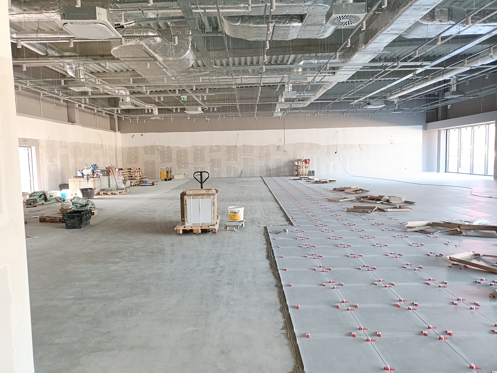 Układanie płytek podłogowych w dużym, surowym wnętrzu. Widoczne płytki z czerwonymi krzyżykami dystansowymi, palety z materiałami i sufit z instalacjami.
