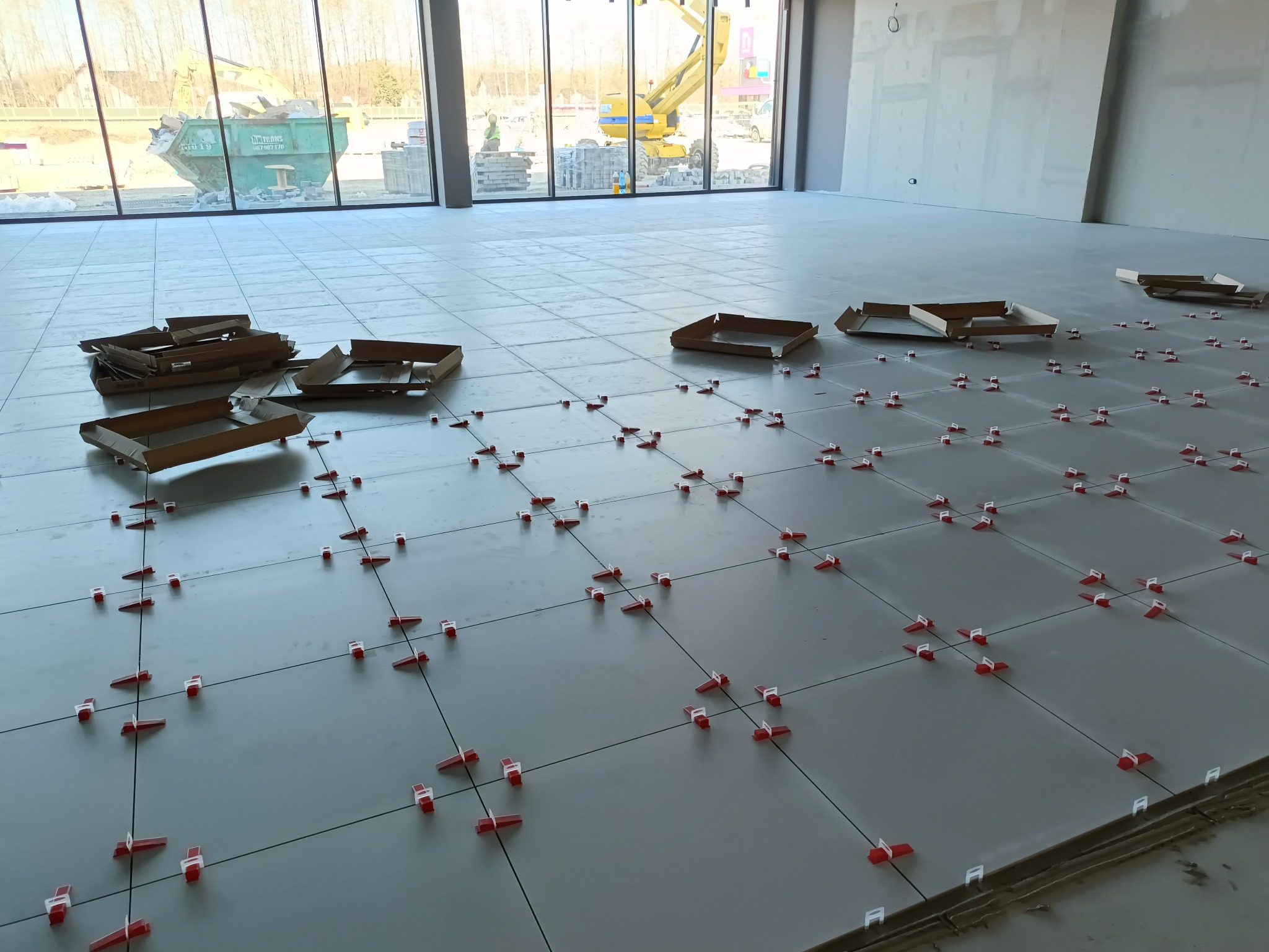 Układanie szarych płytek podłogowych z użyciem czerwonych klipsów poziomujących. W tle okno z widokiem na kontener i sprzęt budowlany. Kartony po płytkach na podłodze.