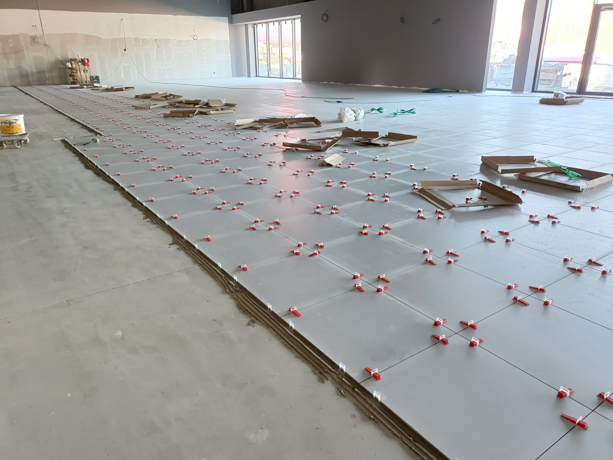 Układanie jasnych płytek podłogowych z użyciem czerwonych klipsów poziomujących w przestronnym, surowym wnętrzu. Widoczne narzędzia i kartony po płytkach.