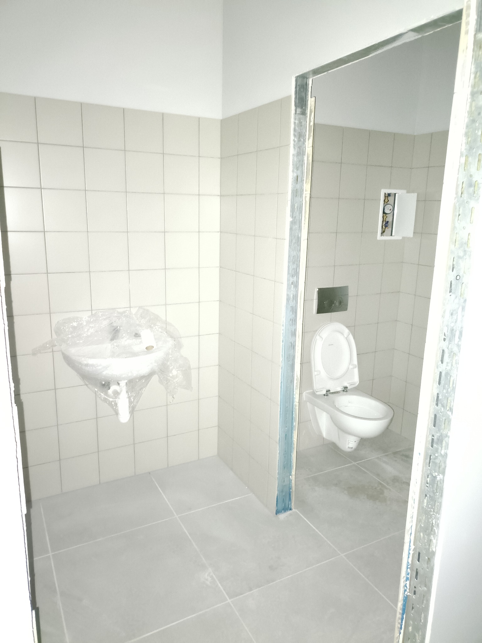 Wykończona łazienka w jasnych płytkach z umywalką pod folią ochronną i toaletą w głębi. Widoczne profile metalowe przy wejściu. Minimalistyczny design.