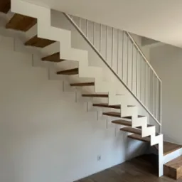 Nowoczesne schody z białymi metalowymi policzkami i drewnianymi stopniami, widoczne z boku, z minimalistyczną białą balustradą i drewnianym podestem startowym.