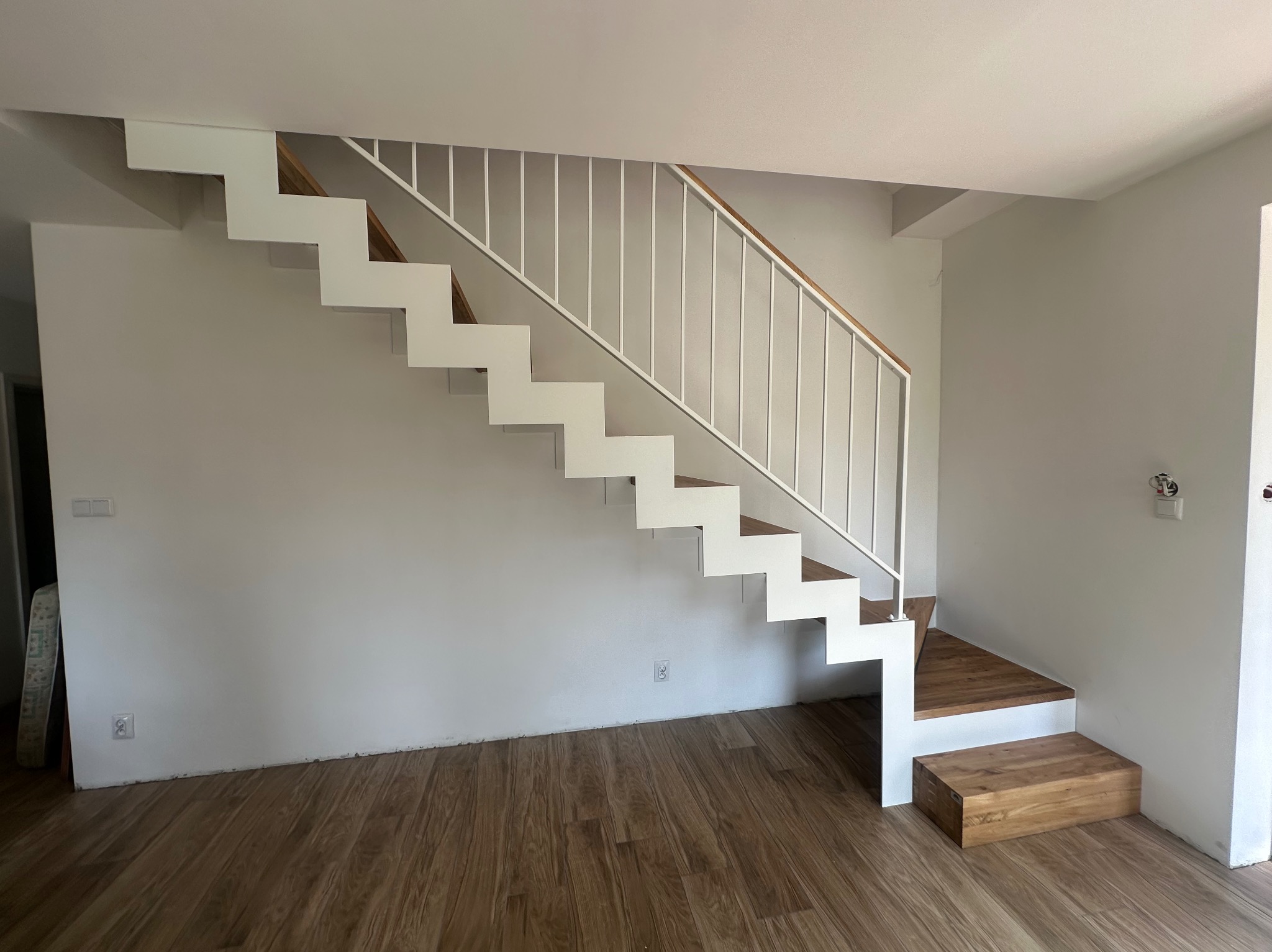 Nowoczesne schody z białymi metalowymi barierkami i drewnianymi stopniami, widok z dołu na jasnym tle ściany i drewnianej podłogi.
