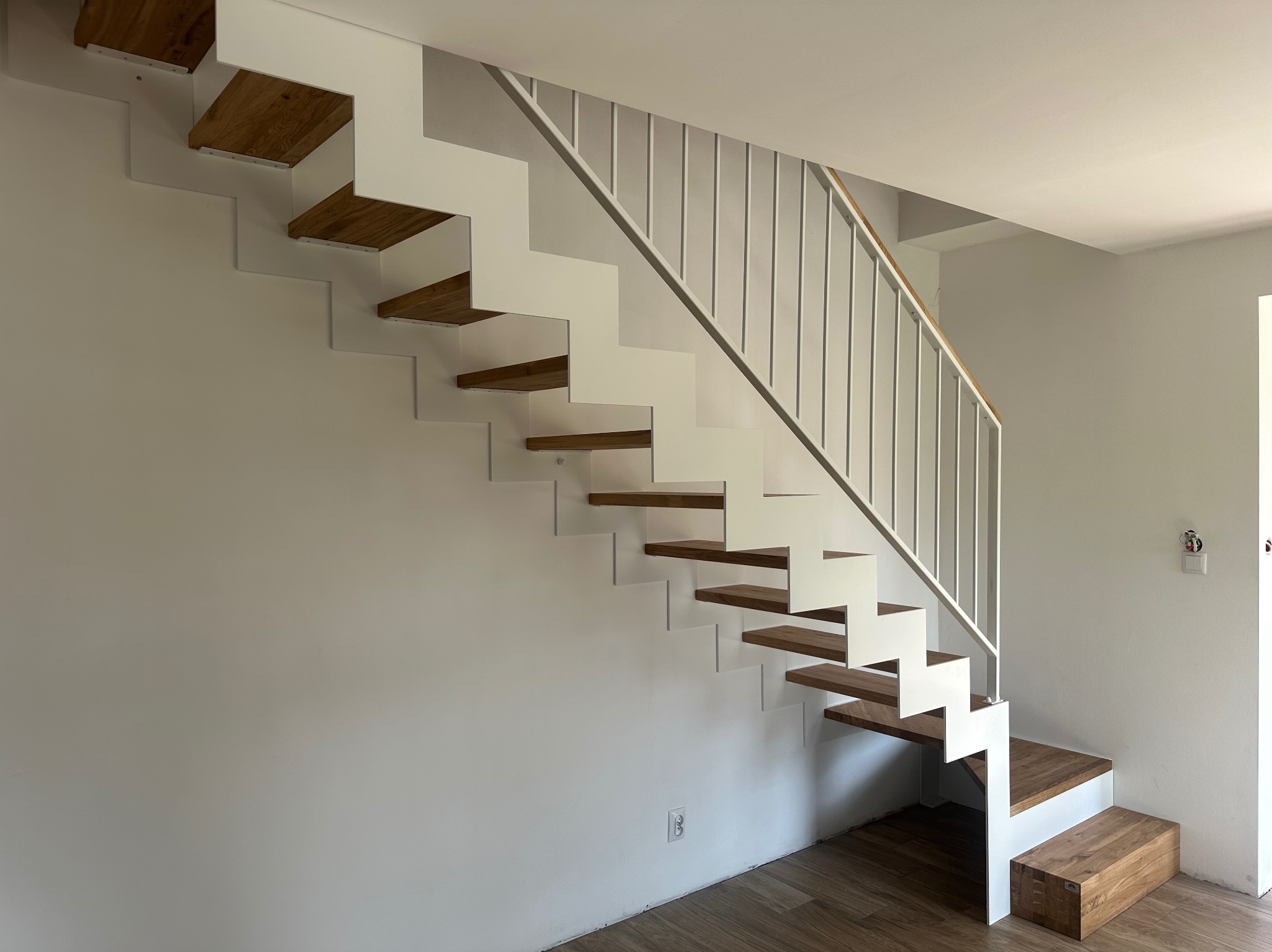Nowoczesne schody z białymi metalowymi policzkami i drewnianymi stopniami, widoczne z boku, z minimalistyczną białą balustradą i drewnianym podestem startowym.