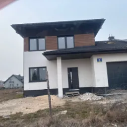 Nowoczesny dom z dwuspadowym dachem, wykończony drewnem i białym tynkiem, z czarnymi oknami. Widoczne resztki materiałów budowlanych. Numer domu '3' na elewacji.