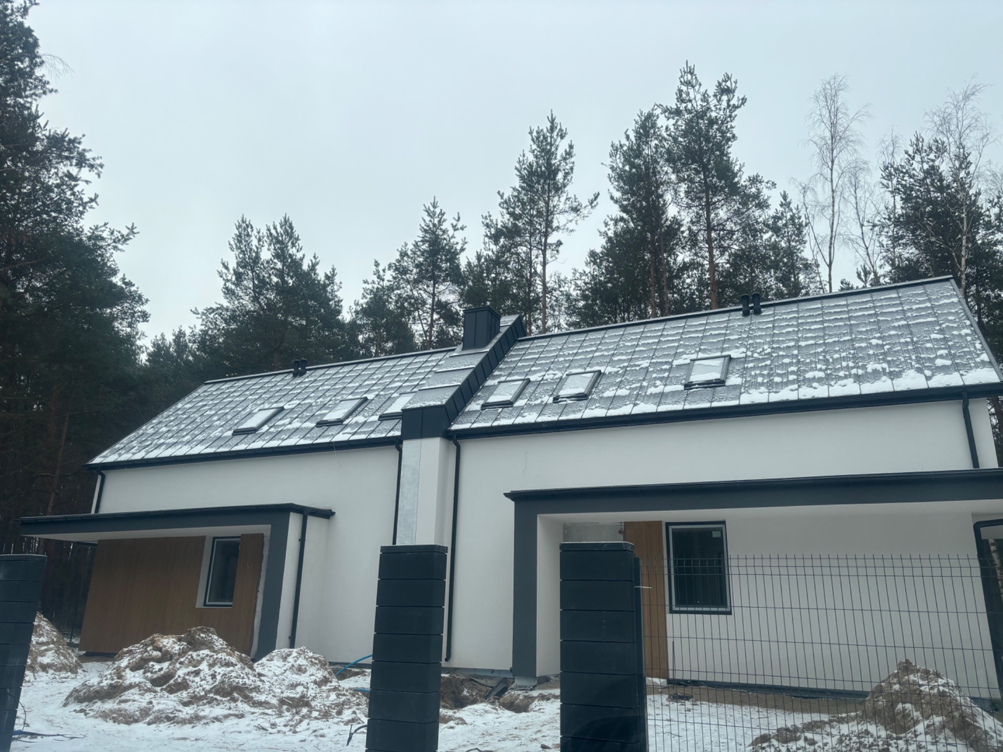 Nowoczesny dom z białymi ścianami i ciemną stolarką okienną, pokryty śniegiem, otoczony lasem. Widoczny fragment ogrodzenia i sterta ziemi.