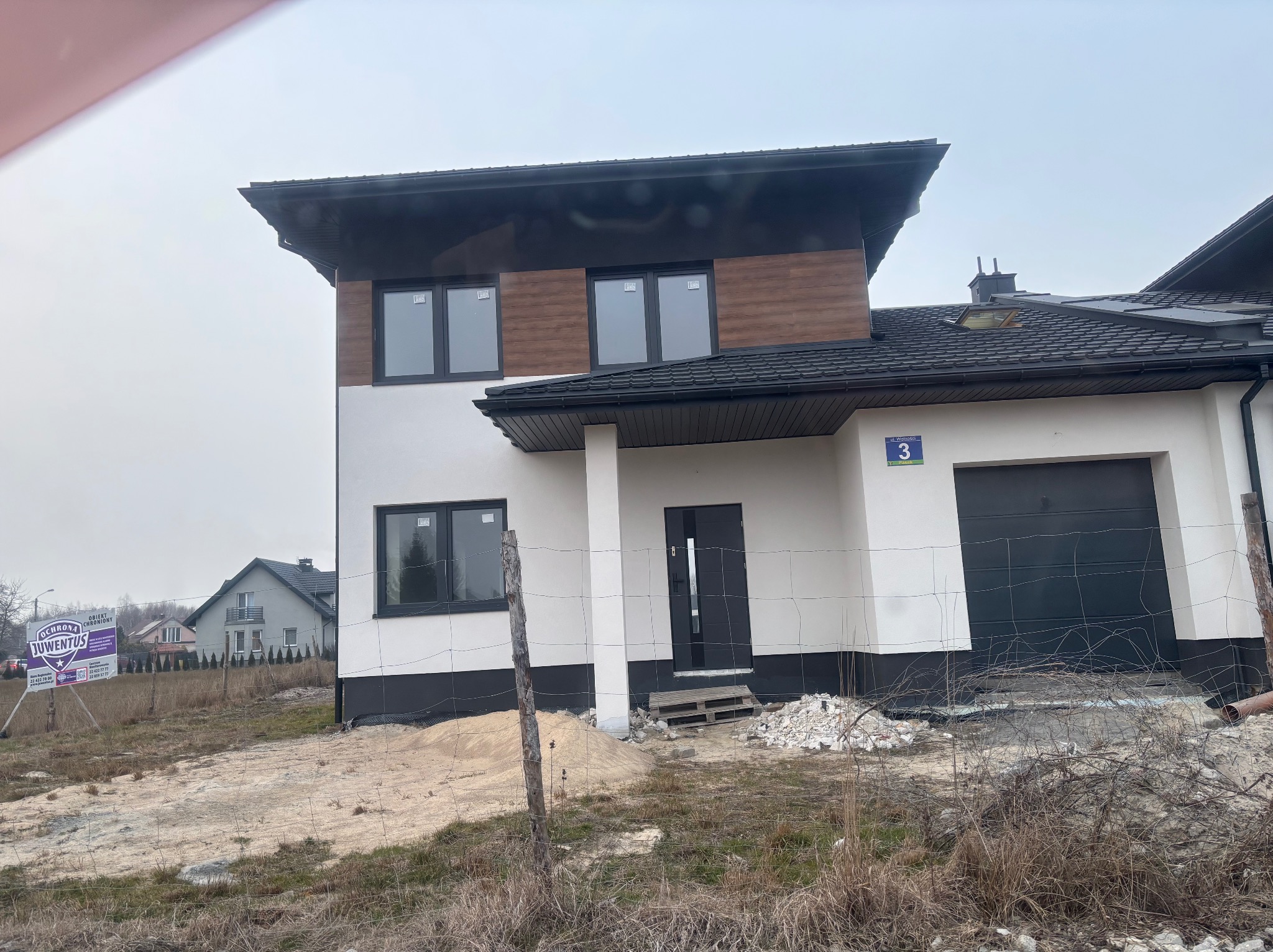 Nowoczesny dom z dwuspadowym dachem, wykończony drewnem i białym tynkiem, z czarnymi oknami. Widoczne resztki materiałów budowlanych. Numer domu '3' na elewacji.