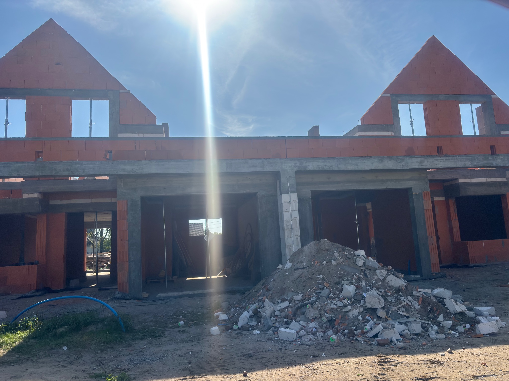 Surowa konstrukcja bliźniaka z czerwonej cegły, w trakcie budowy. Widoczne okna i zarys dachu, w tle błękitne niebo i ostre światło słoneczne. Na pierwszym planie gruz.