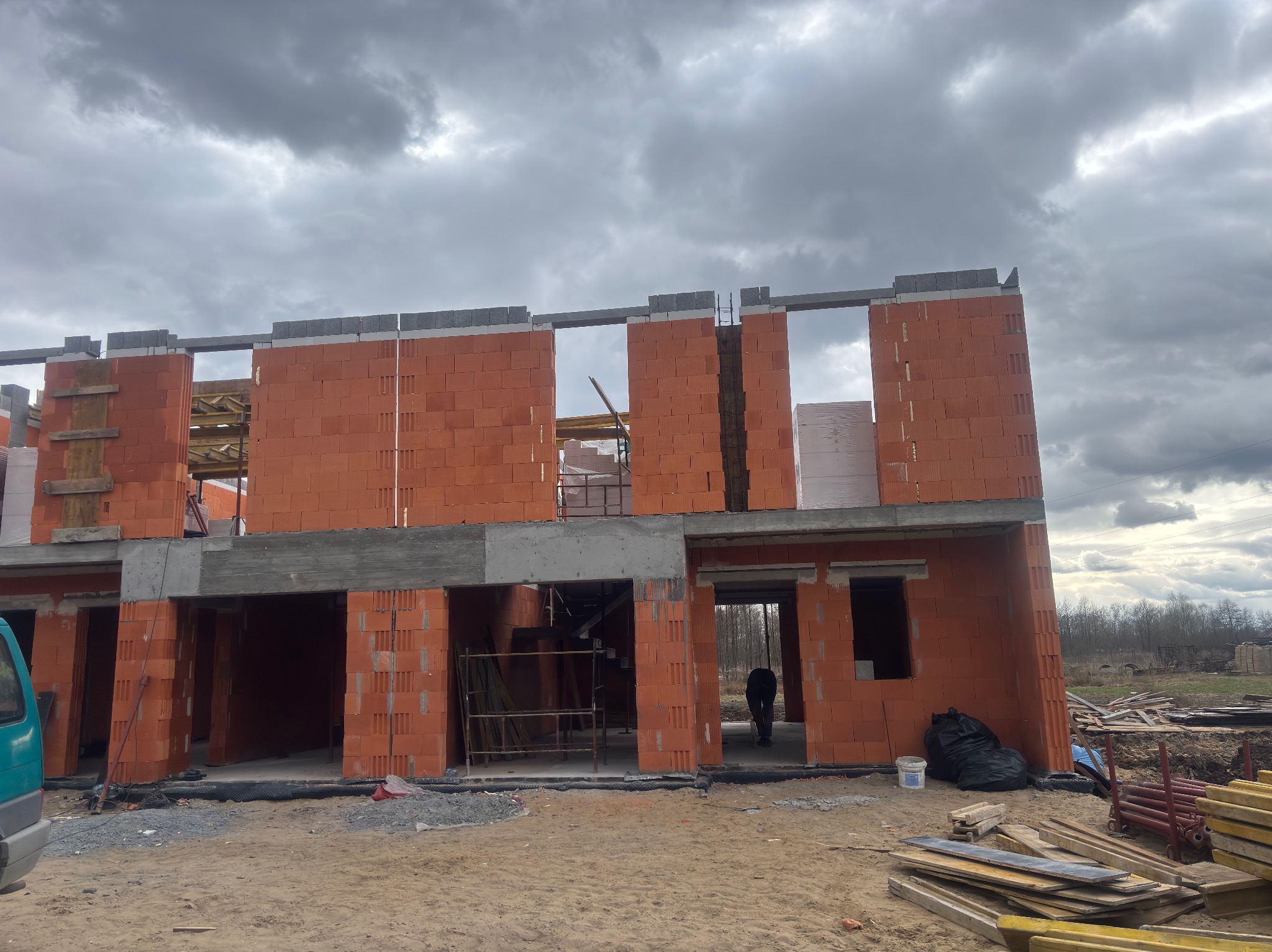 Wznoszenie ścian domu z czerwonej cegły ceramicznej. Widoczne nadproża, elementy konstrukcyjne i szary, pochmurny krajobraz w tle. Budowa w trakcie realizacji.