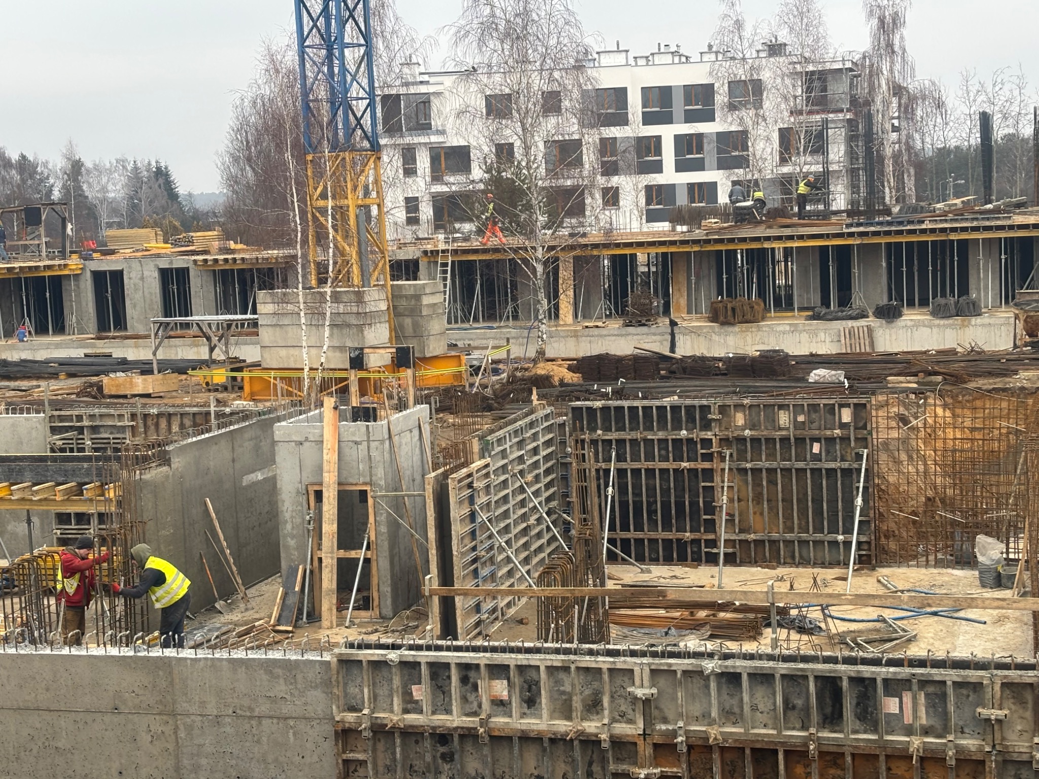 Budowa wielorodzinnego budynku w trakcie wznoszenia. Widoczne szalunki, zbrojenia, betonowe ściany i pracujący robotnicy w odblaskowych kamizelkach. Kran budowlany w tle.