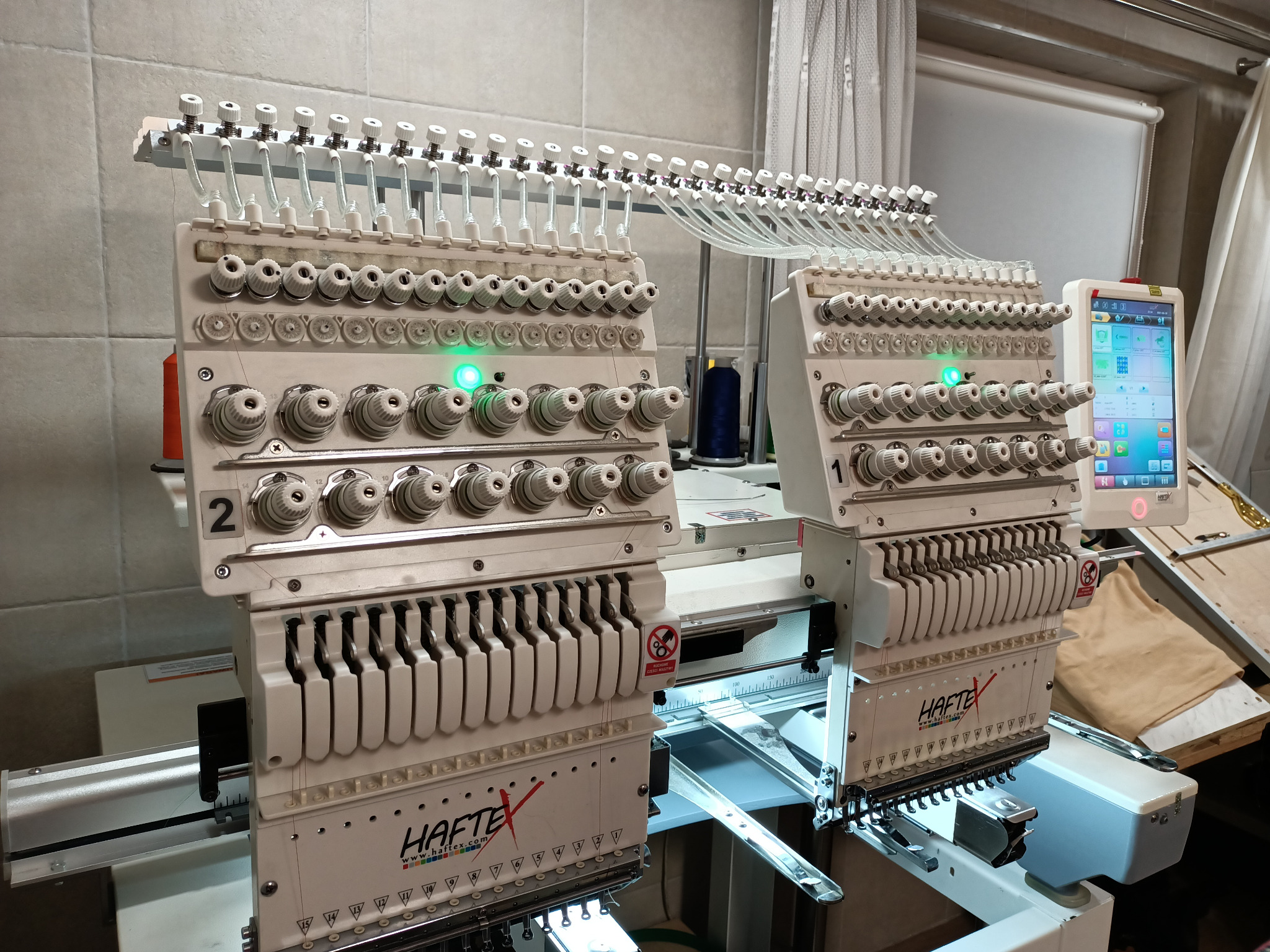 Dwie białe, wielogłowicowe maszyny do szycia przemysłowego z nawleczonymi nićmi, widoczne detale konstrukcyjne i panel sterowania z ekranem dotykowym.