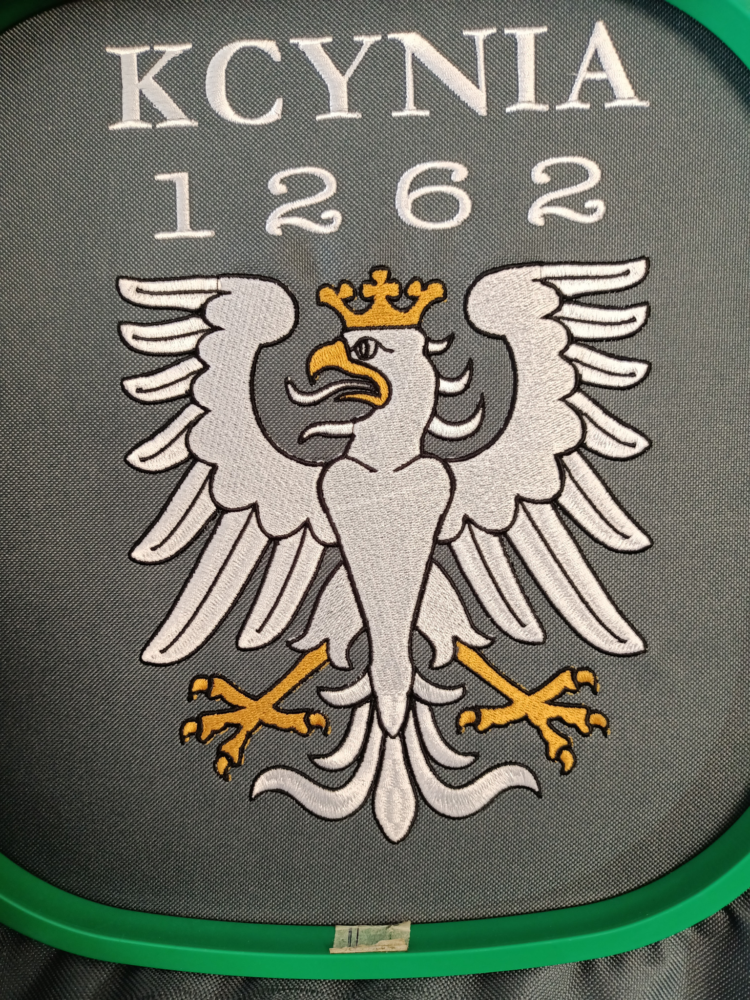 Herb miasta Kcynia z datą 1262, wykonany techniką haftu na szarym tle, umieszczony w zielonej ramce.