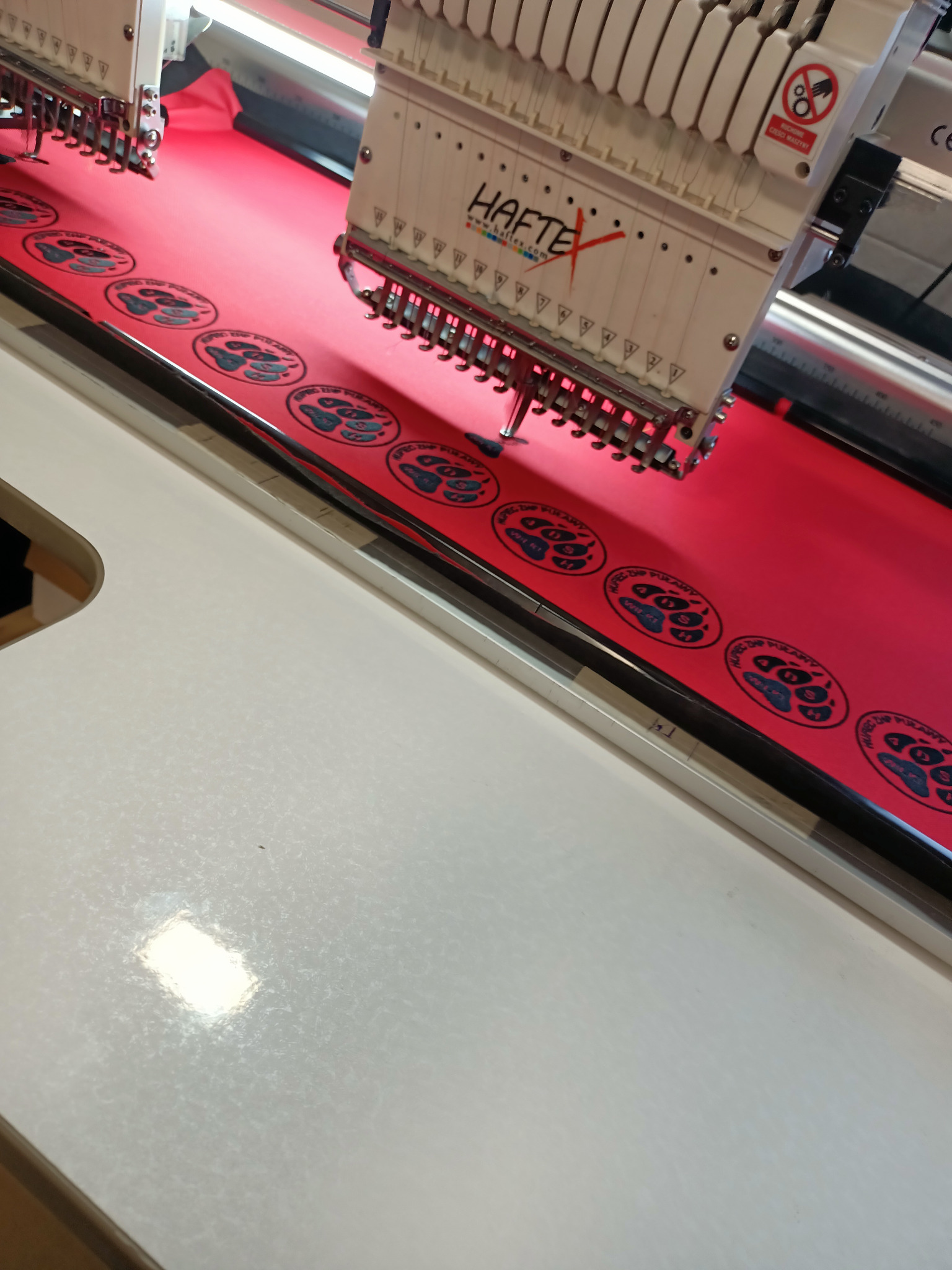 Automat szwalniczy w trakcie nanoszenia haftu z logo na czerwony materiał, widoczne igły i gotowe wzory z napisem Spec Chip Prawy.