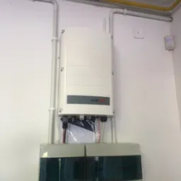 Biały falownik SolarEdge zamontowany na ścianie z szarą skrzynką bezpieczników ABB poniżej, widoczne rurki instalacyjne i czujnik ruchu w tle.