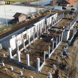 Nazarii Tesliuk TIN - Widok z lotu ptaka na fundamenty budynku z betonowymi słupami i ścianami, otoczone ziemią i placem budowy. W tle widoczny teren rekreacyjny.