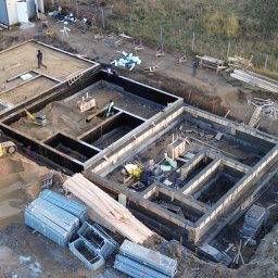 Nazarii Tesliuk TIN - Zalane fundamenty domu z szalunkiem, widok z góry. Na pierwszym planie stosy metalowych elementów szalunkowych i żółta koparka. Prace ziemne w toku.