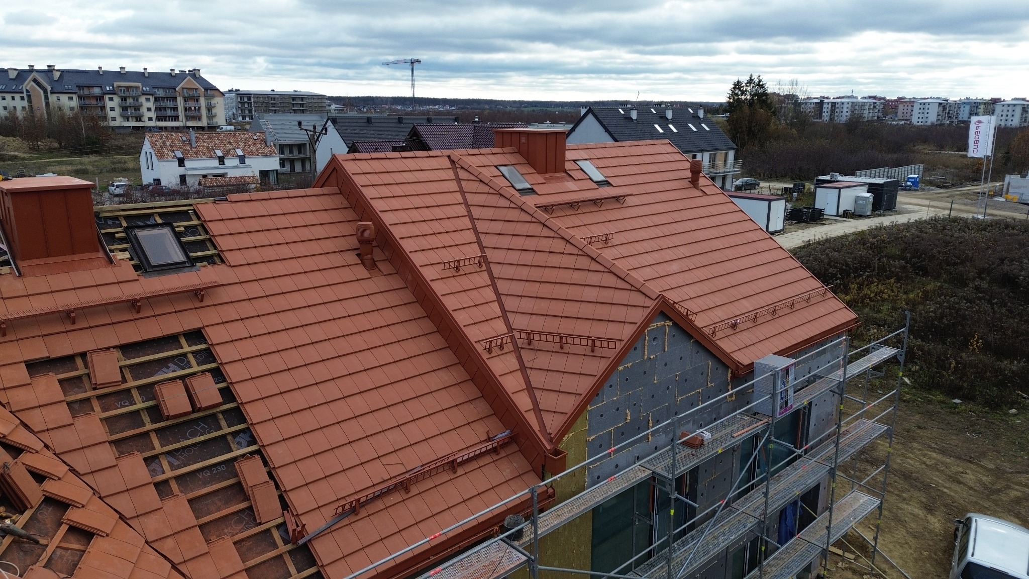 Ceglasty dach z lukarnami w trakcie montażu, widok z góry. Część dachu bez pokrycia, widoczne elementy konstrukcyjne. Budynek w trakcie budowy, rusztowania.