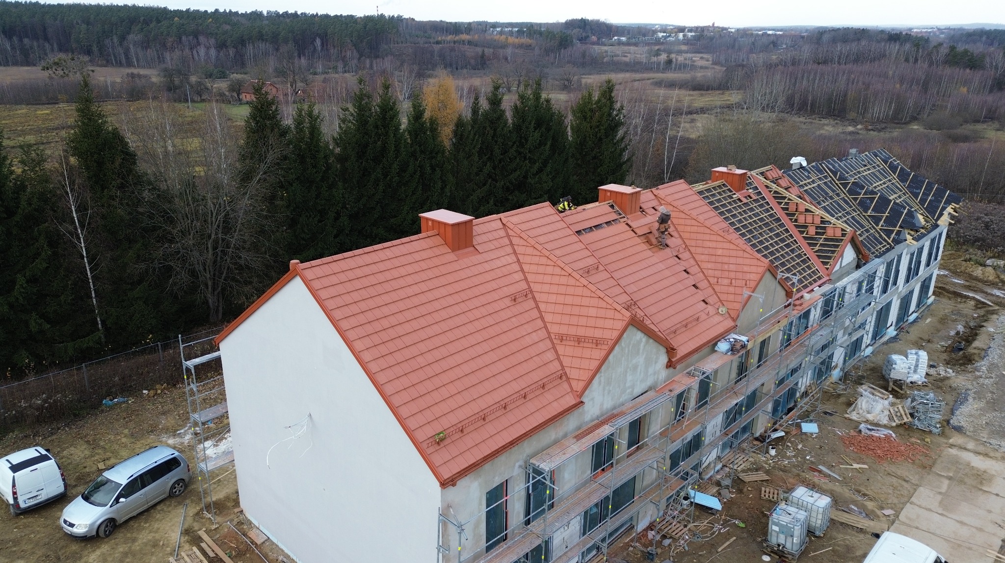 Wielorodzinny budynek w trakcie budowy dachu z czerwoną dachówką. Część dachu pokryta czarną membraną. Widoczne rusztowania i pracownicy na dachu. Ujęcie z drona.