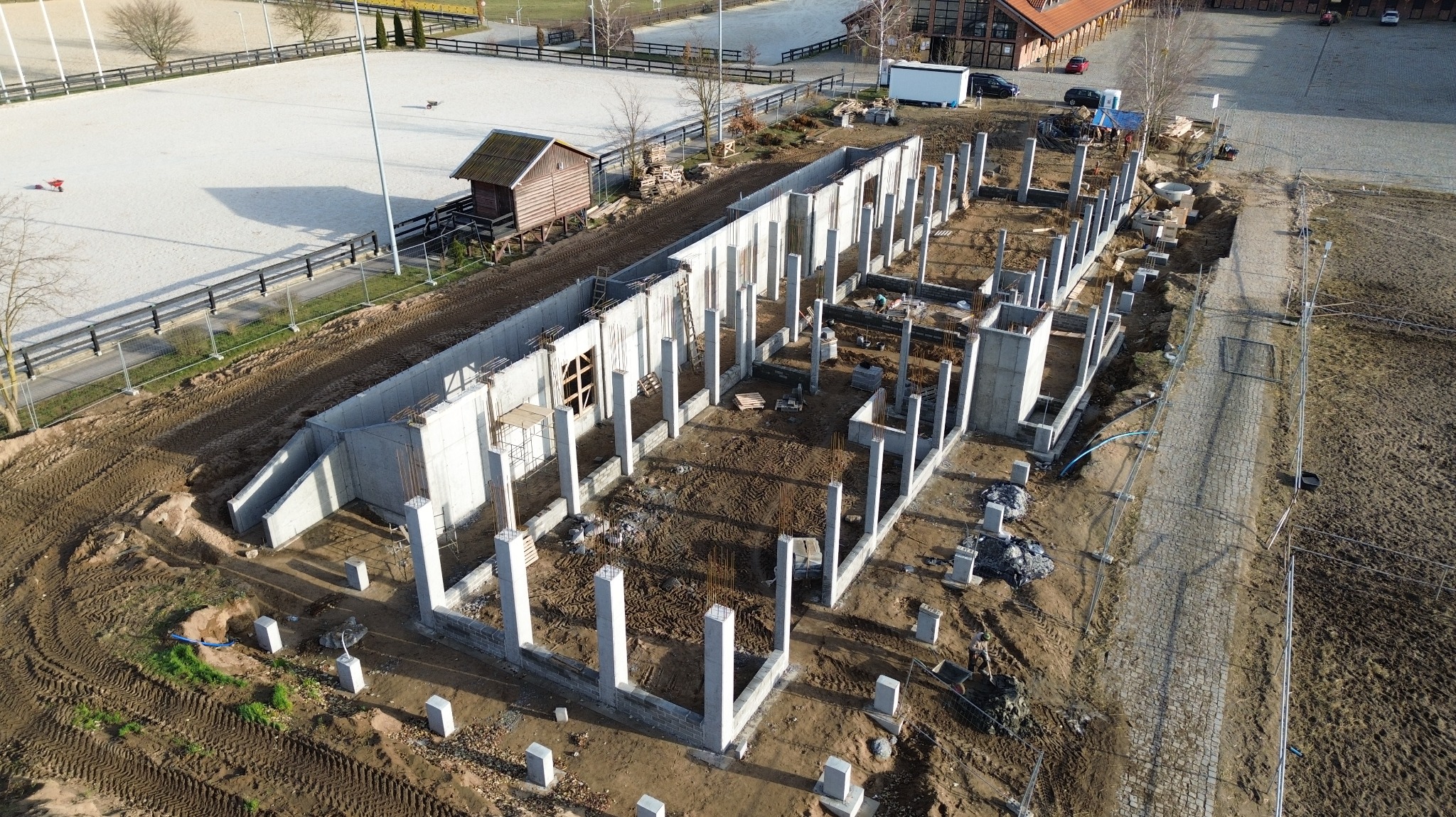 Widok z lotu ptaka na fundamenty budynku z betonowymi słupami i ścianami, otoczone ziemią i placem budowy. W tle widoczny teren rekreacyjny.