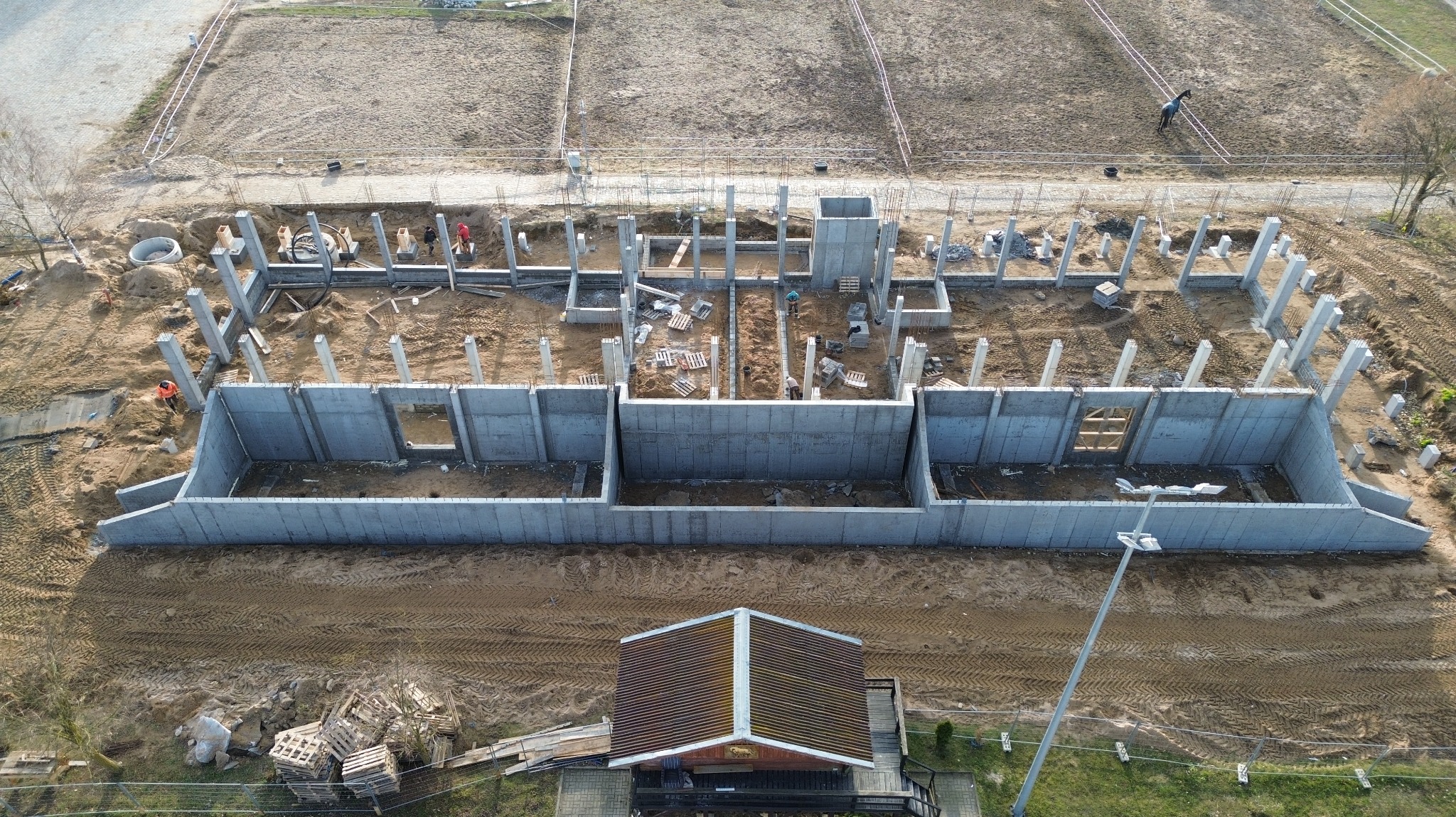 Budowa domu w toku: widok z góry na fundamenty i słupy konstrukcyjne. Na pierwszym planie budynek z brązowym dachem. Prace budowlane w słoneczny dzień.