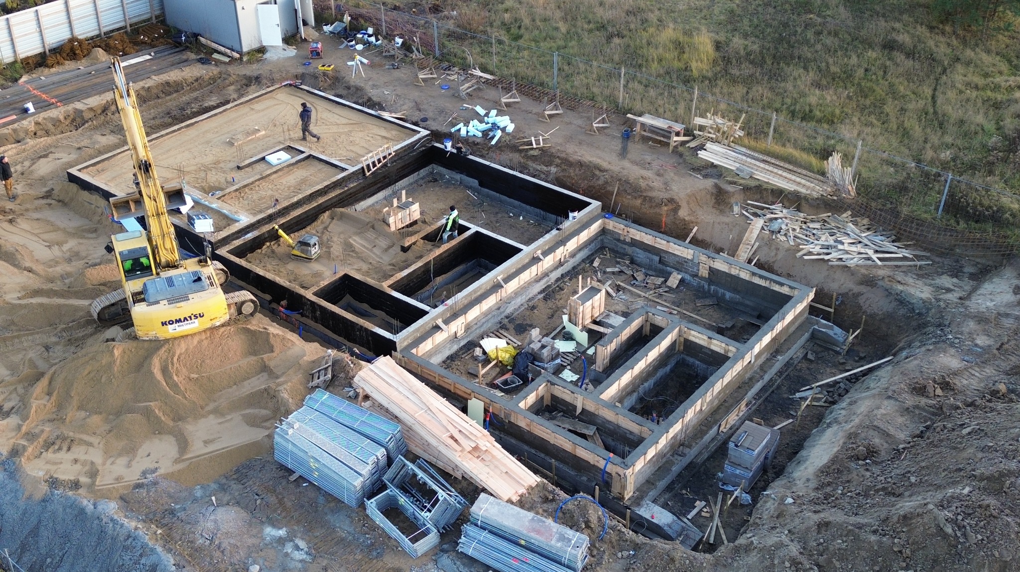 Zalane fundamenty domu z szalunkiem, widok z góry. Na pierwszym planie stosy metalowych elementów szalunkowych i żółta koparka. Prace ziemne w toku.