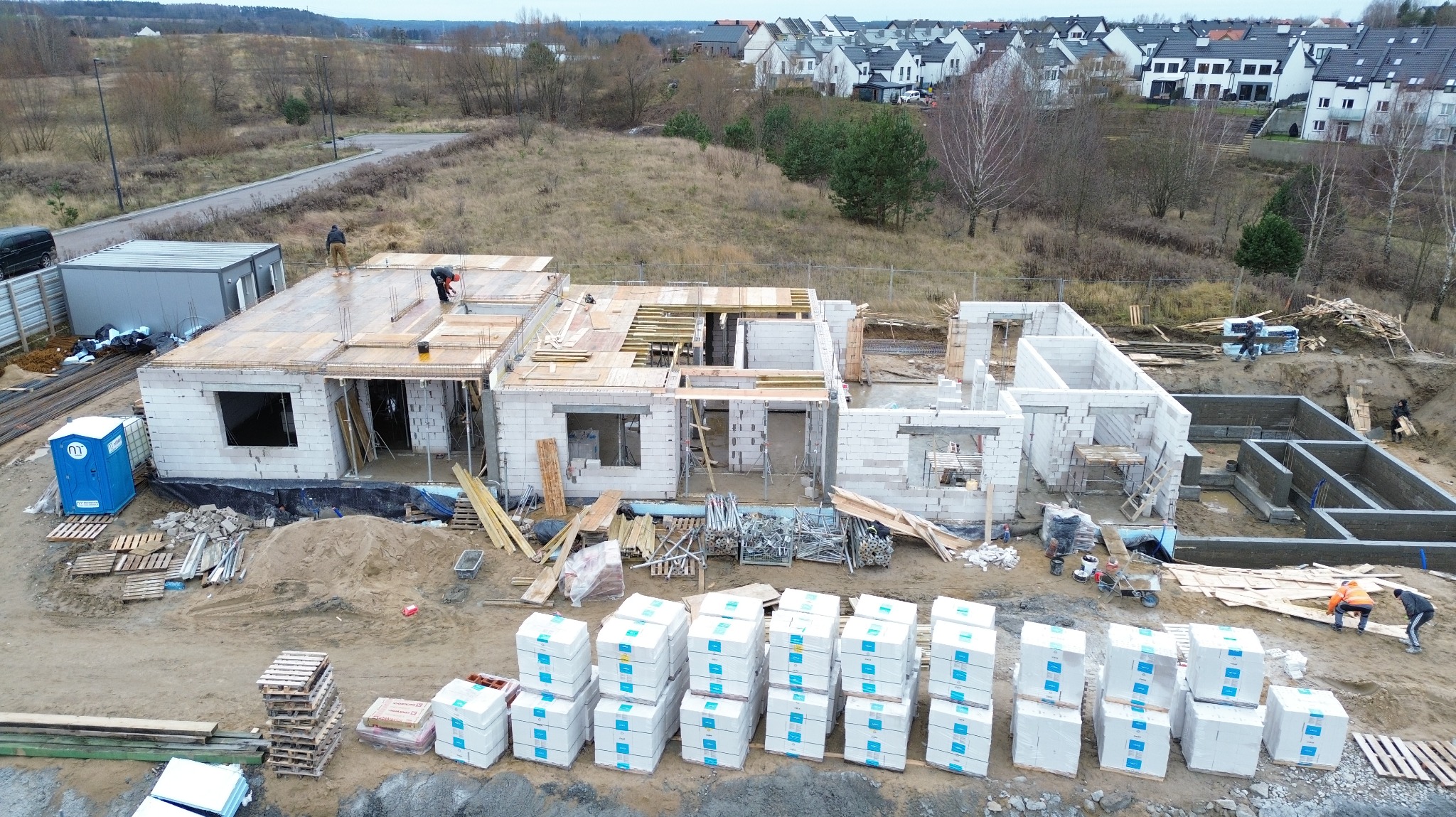 Budowa domu z betonu komórkowego w trakcie realizacji. Widok z góry na fundamenty, ściany i strop. Na dachu pracują robotnicy. W tle zabudowa jednorodzinna.
