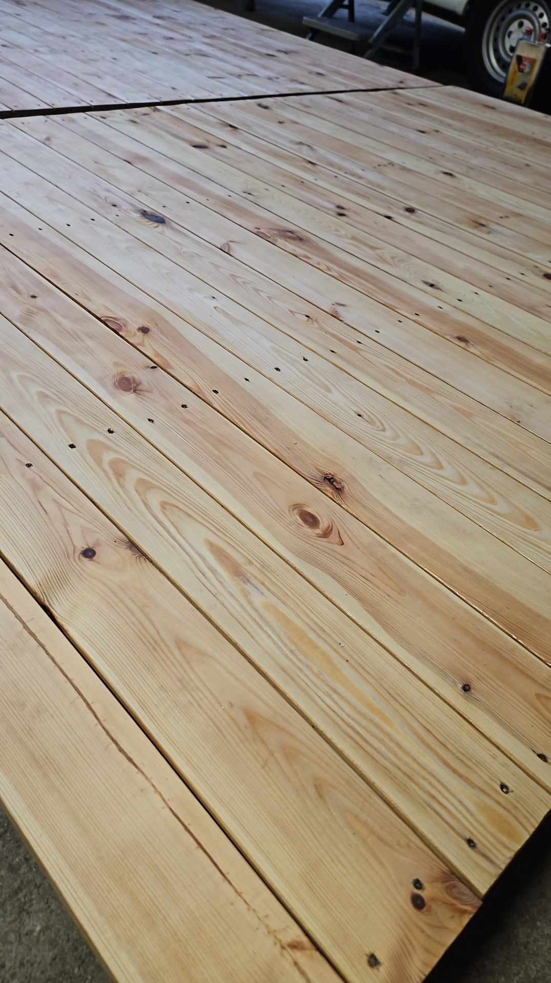 Jasne, świeżo ułożone deski tarasowe z widocznymi sękami i wkrętami, tworzące regularny wzór. W tle widoczny fragment samochodu.
