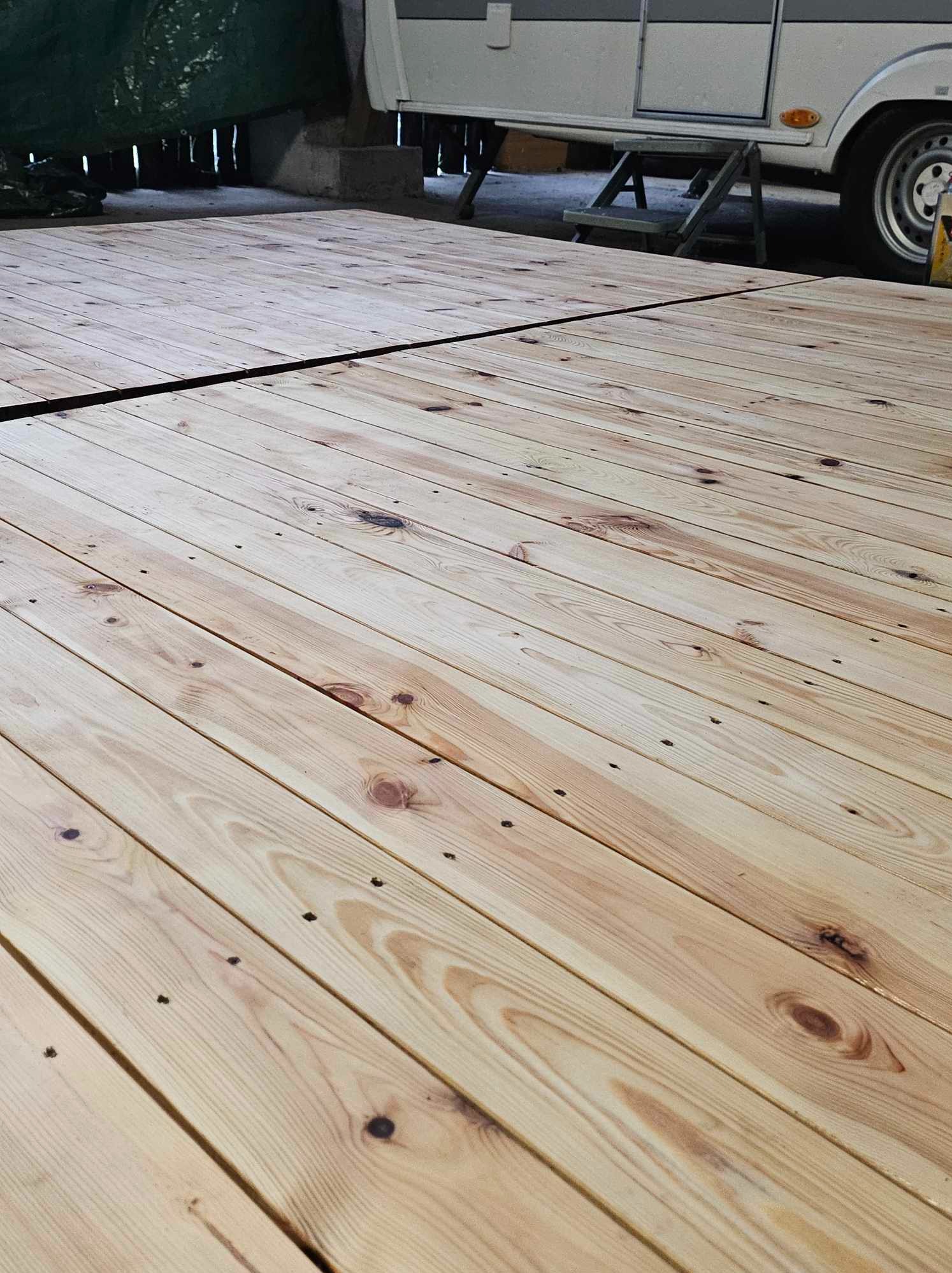 Nowo ułożona, jasna drewniana podłoga z widocznymi sękami i drobnymi otworami po wkrętach, częściowo zadaszona, z przyczepą kempingową w tle.