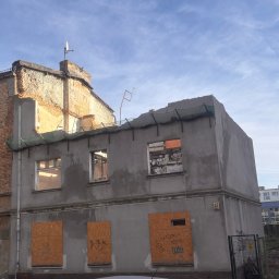 Dominik Małecki - Budynek w trakcie rozbiórki z zabitymi deskami oknami, widoczny fragment samochodu i pracownik w kasku. Częściowo zdemontowany dach i elewacja. Numer budynku 15.