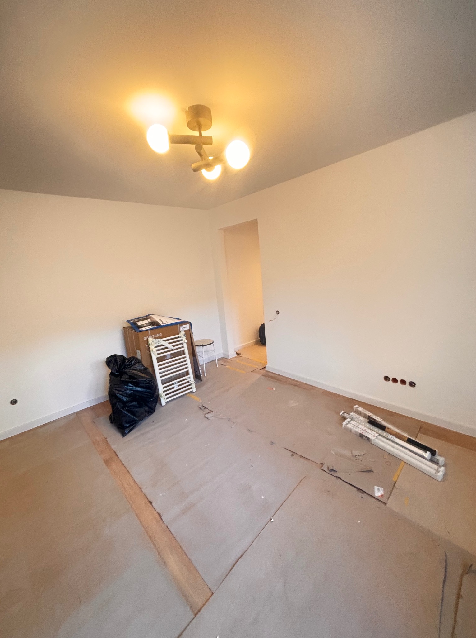 Pokój w trakcie remontu: gołe ściany, podłoga przygotowana pod panele, widoczne materiały budowlane i worek na śmieci. Oświetlenie sufitowe włączone.