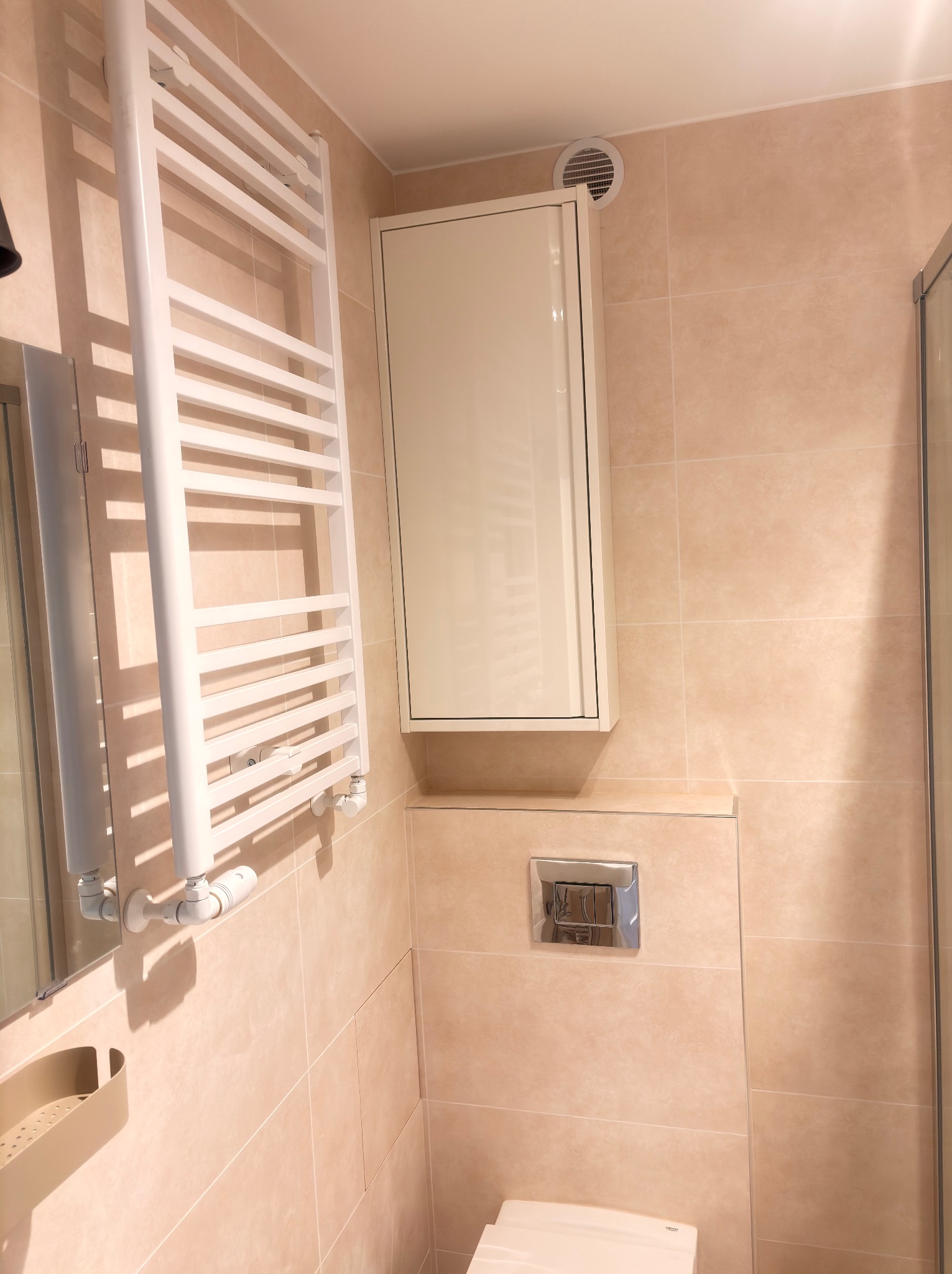 Wykończona łazienka w odcieniach beżu w Poznaniu. Widoczny grzejnik drabinkowy, szafka wisząca i toaleta. Minimalistyczny design, płytki ceramiczne na ścianach.