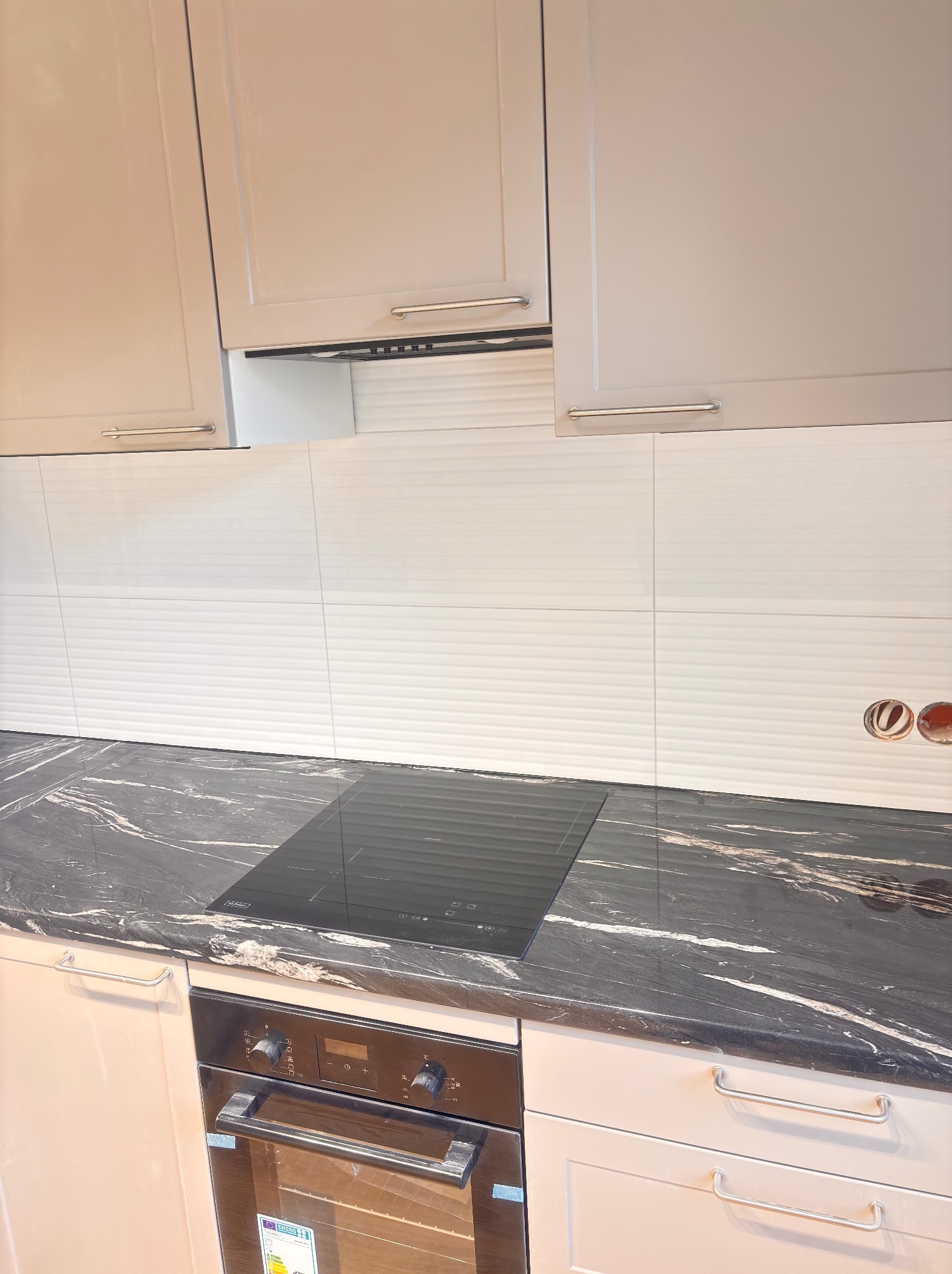 Wykończona kuchnia z czarnym blatem, płytą indukcyjną i białymi, poziomymi płytkami na ścianie. Widoczne szafki górne i piekarnik w zabudowie.