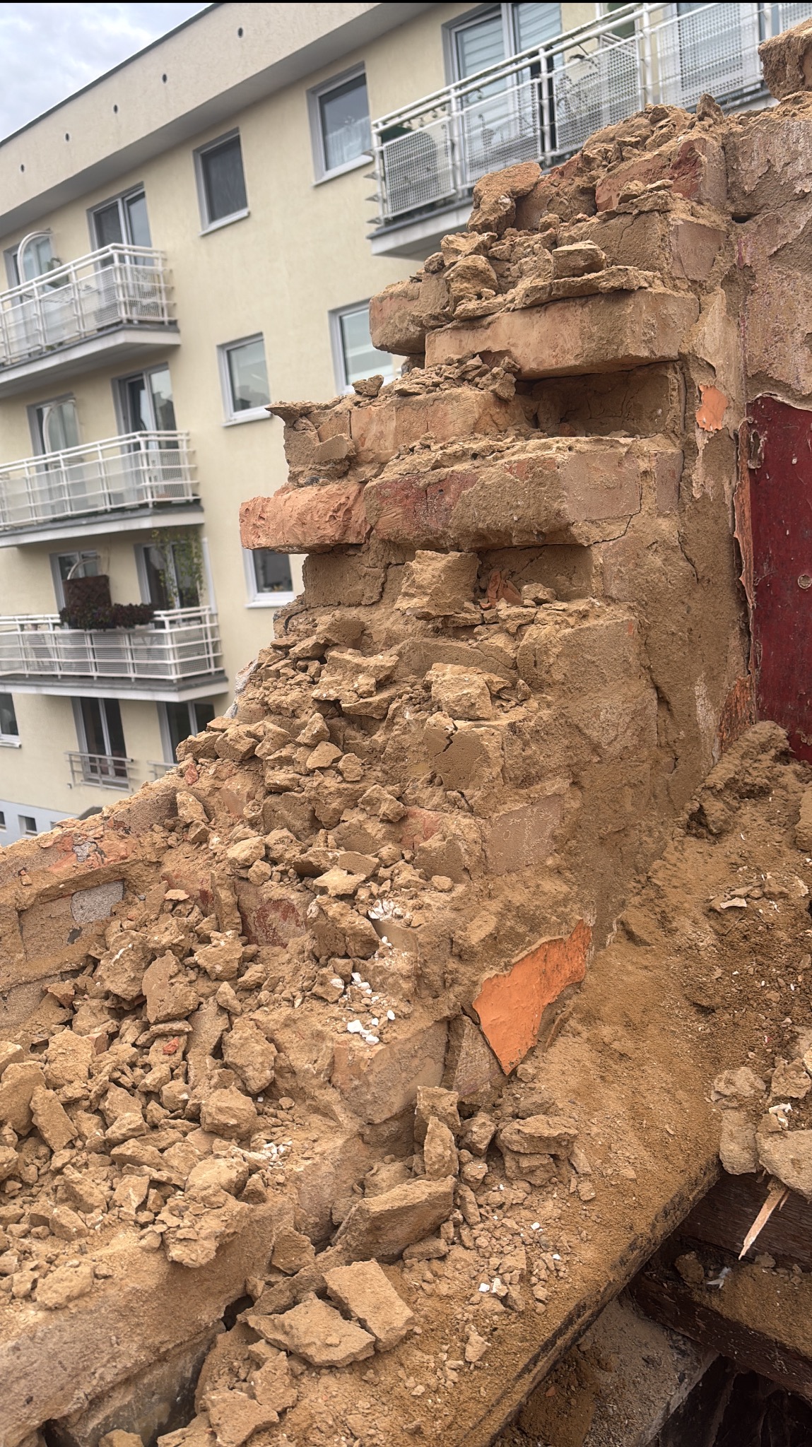 Fragment zniszczonej, ceglanej ściany w trakcie rozbiórki, z widocznymi gruzami i fragmentami cegieł na tle budynku mieszkalnego.