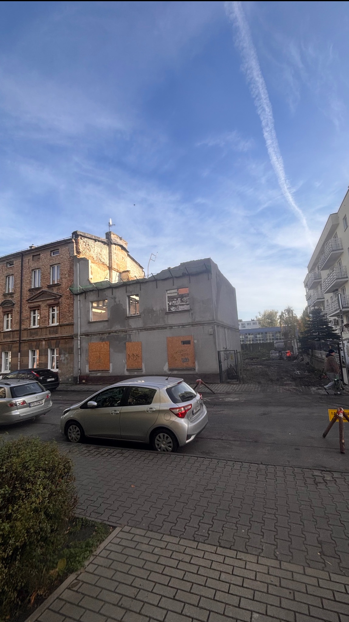Budynek po częściowej rozbiórce, z zabitymi deskami oknami, widoczny z ulicy z zaparkowanymi samochodami i fragmentem chodnika z kostki.