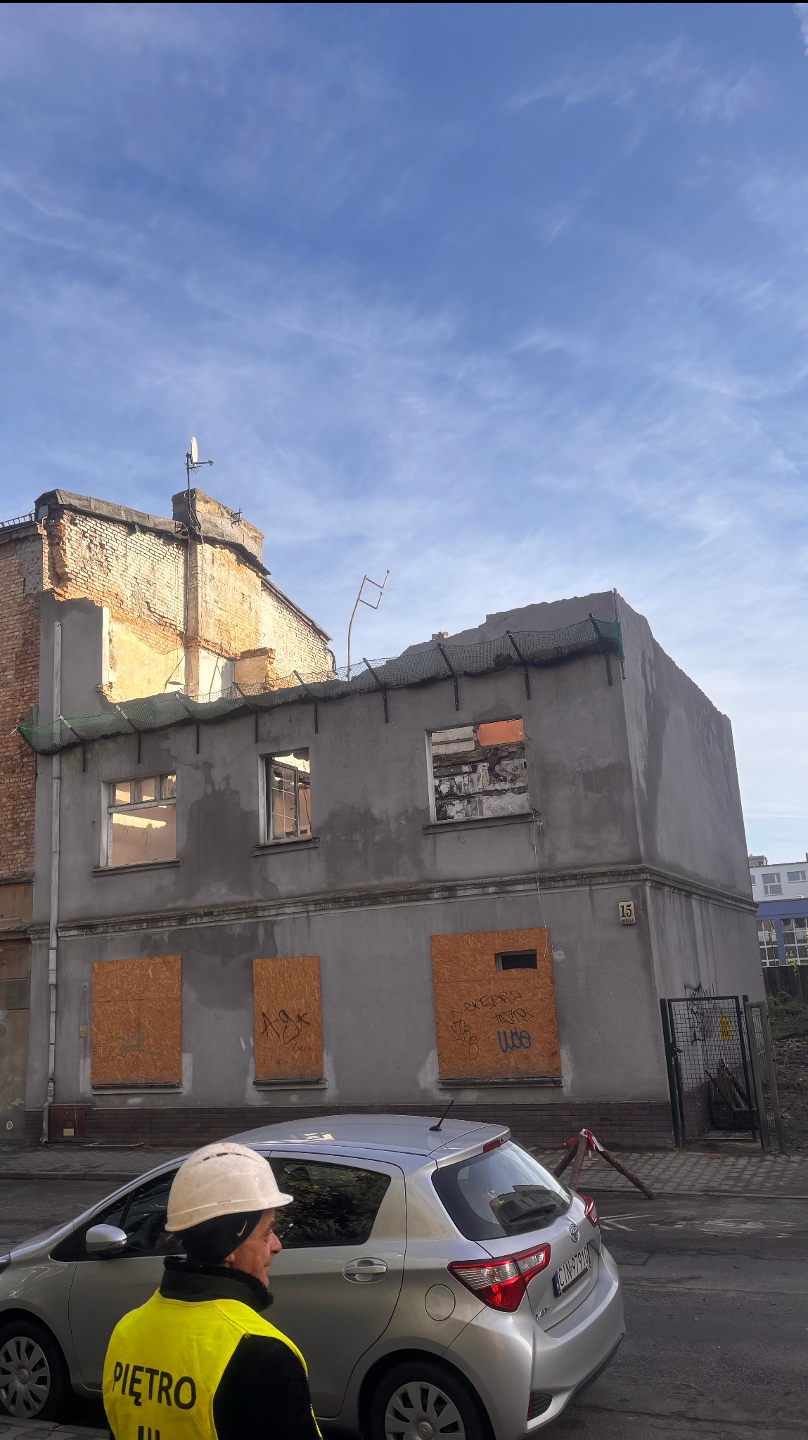 Budynek w trakcie rozbiórki z zabitymi deskami oknami, widoczny fragment samochodu i pracownik w kasku. Częściowo zdemontowany dach i elewacja. Numer budynku 15.