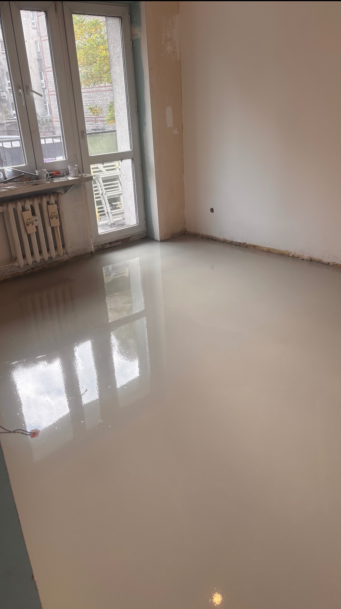 Wylana, gładka posadzka samopoziomująca w pokoju z oknem i grzejnikiem. Ściany przygotowane do malowania. Widoczne ślady po remoncie.