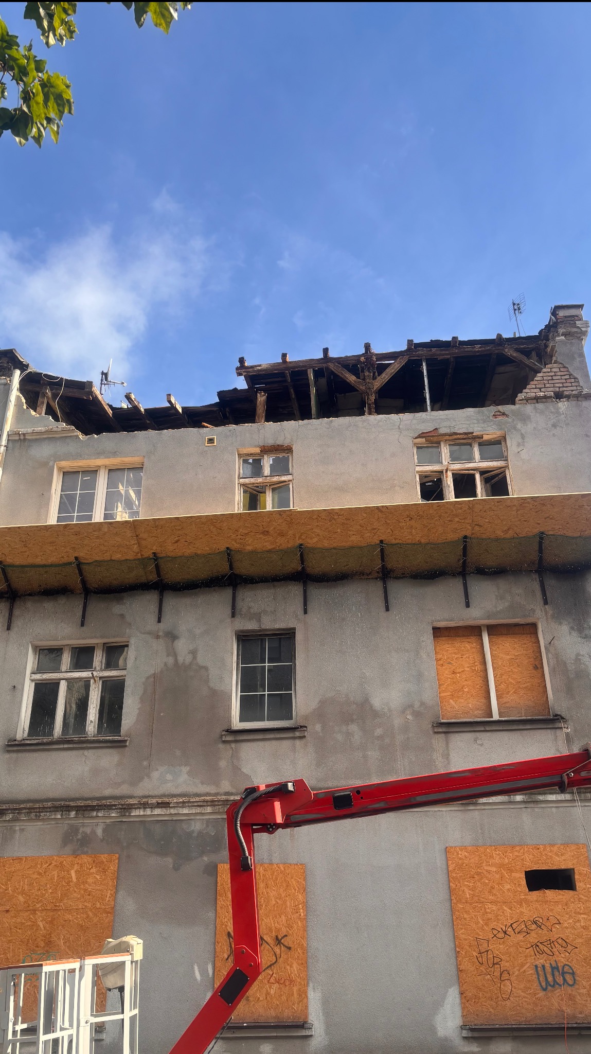 Budynek w trakcie rozbiórki, z odsłoniętą konstrukcją dachu i zabezpieczonymi oknami płytami OSB. Widoczny fragment podnośnika koszowego.