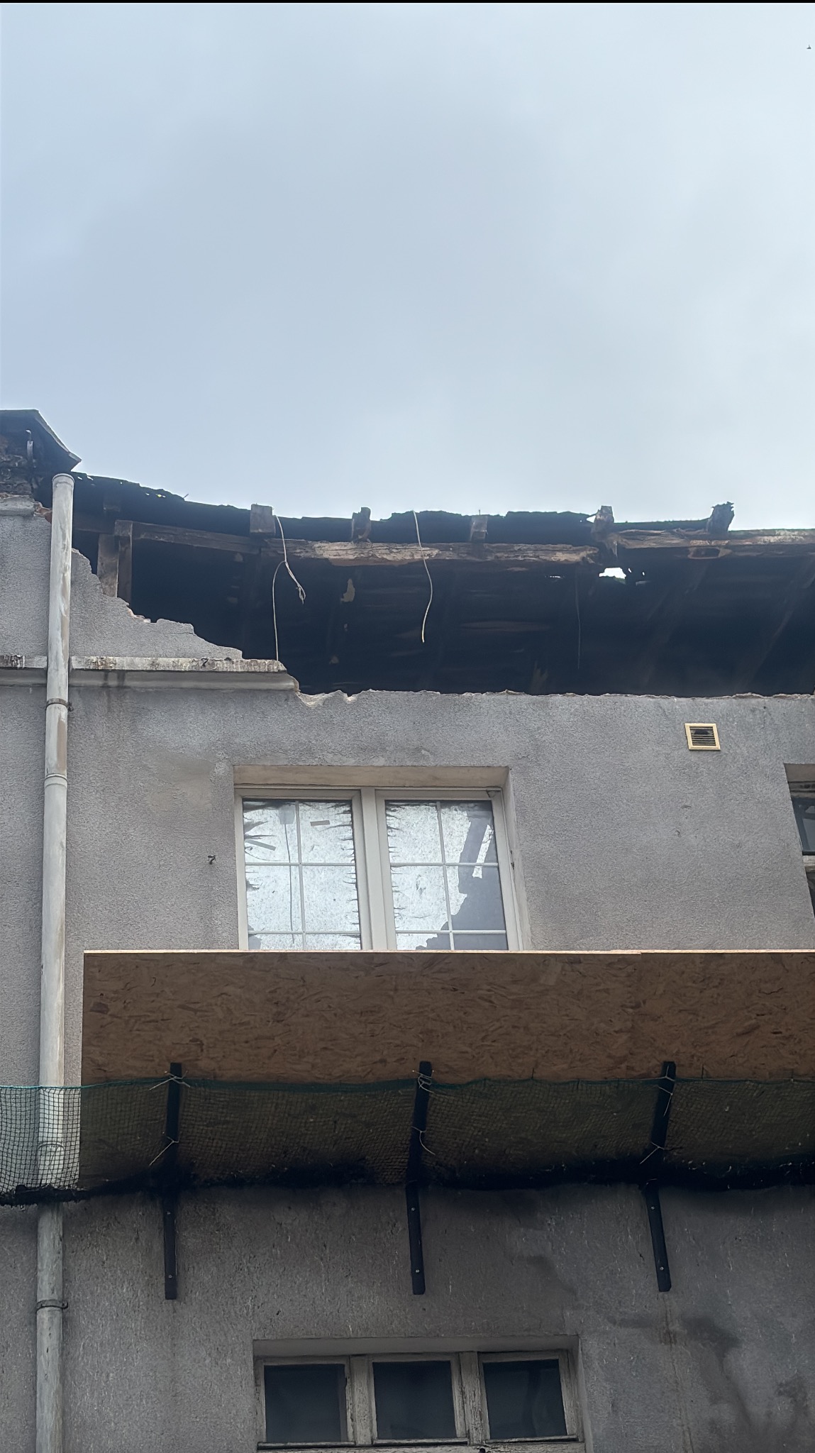 Uszkodzony dach budynku z zabezpieczonym oknem. Widoczne ślady po pożarze i prowizoryczne zabezpieczenia. Szary budynek z widocznymi oknami i elementami konstrukcyjnymi.