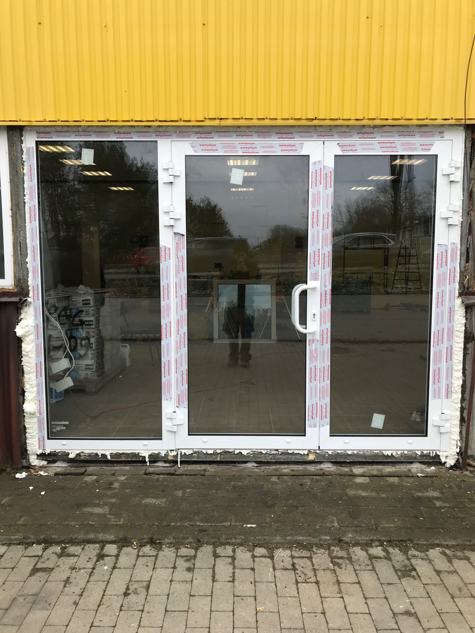 Trzyczęściowe białe drzwi z szybami, widoczne ślady montażu pianką, żółta elewacja budynku w tle, odbicie osoby w szybie.